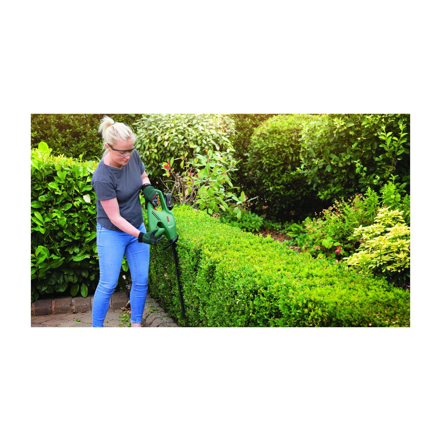Una donna con gli occhiali taglia una siepe verde con un tagliasiepi elettrico verde, con jeans e maglietta grigia, in un giardino.
