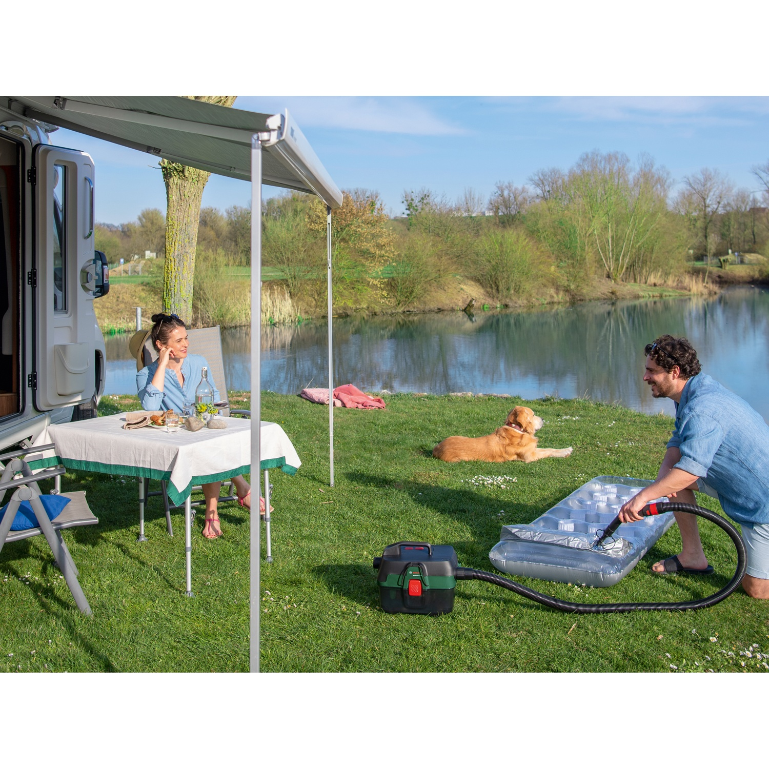 Una coppia fa un picnic in riva al lago. Un uomo gonfia un materasso ad aria. Un cane si rilassa nelle vicinanze. Un camper è visibile.
