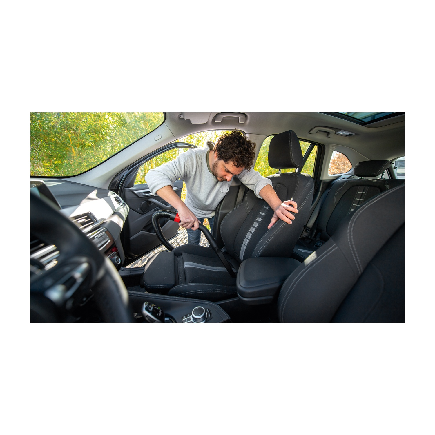 Un uomo aspira l'interno di un'auto. Indossa un maglione grigio. Sono visibili sedili neri e un aspirapolvere nero. Lo sfondo è di alberi verdi.