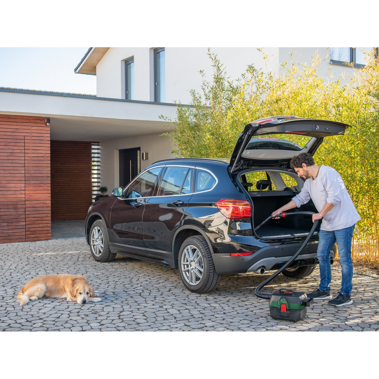 Un uomo aspira il bagagliaio di un'auto nera. Un golden retriever giace sul vialetto pavimentato vicino all'auto.