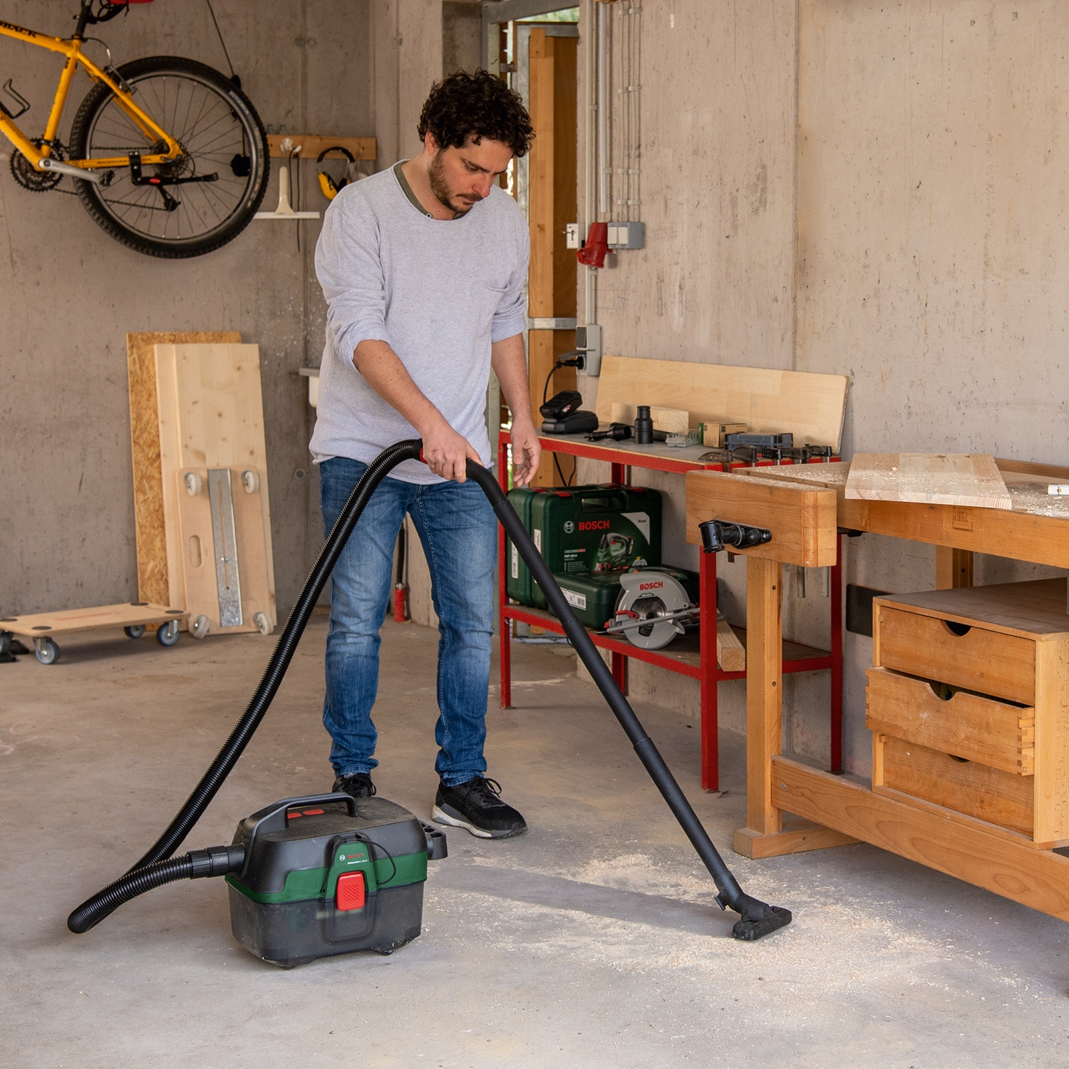 Un uomo aspira il pavimento di un garage. Una bicicletta gialla è appesa al muro. Un banco da lavoro e degli attrezzi sono visibili.