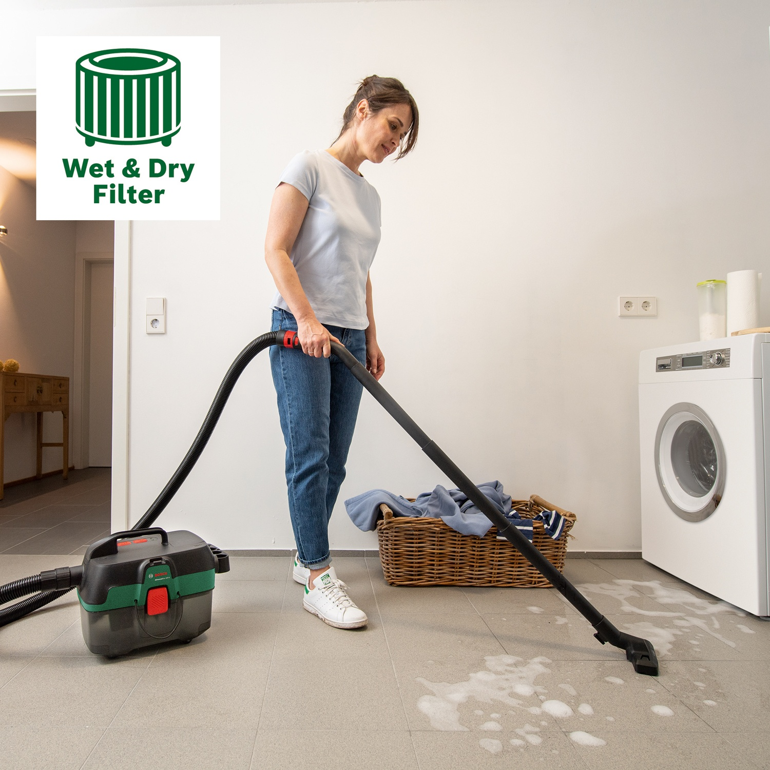 Donna che aspira un pavimento bagnato. Visibili un aspirapolvere per liquidi e solidi, un cesto della biancheria, una lavatrice. Un logo recita 'Wet & Dry Filter'.
