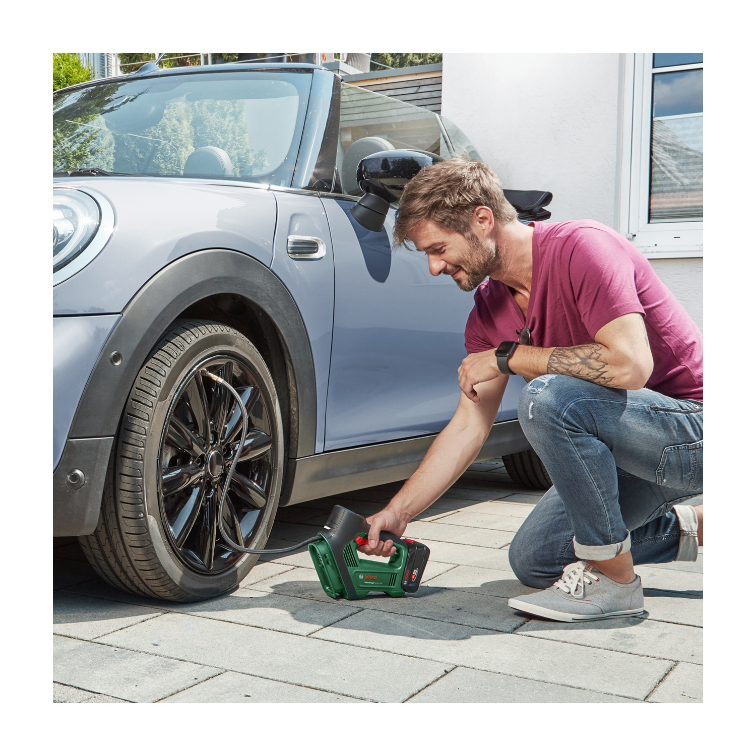 Un uomo è in ginocchio vicino a un'auto grigia, gonfia un pneumatico con uno strumento verde. Indossa una maglietta rosa e jeans. Lo sfondo è una casa.
