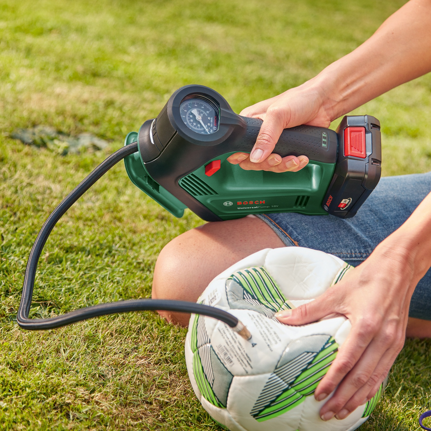 Una persona gonfia un pallone da calcio con una pompa Bosch verde e nera sull'erba. La palla ha strisce verdi. Lo sfondo è erba verde.