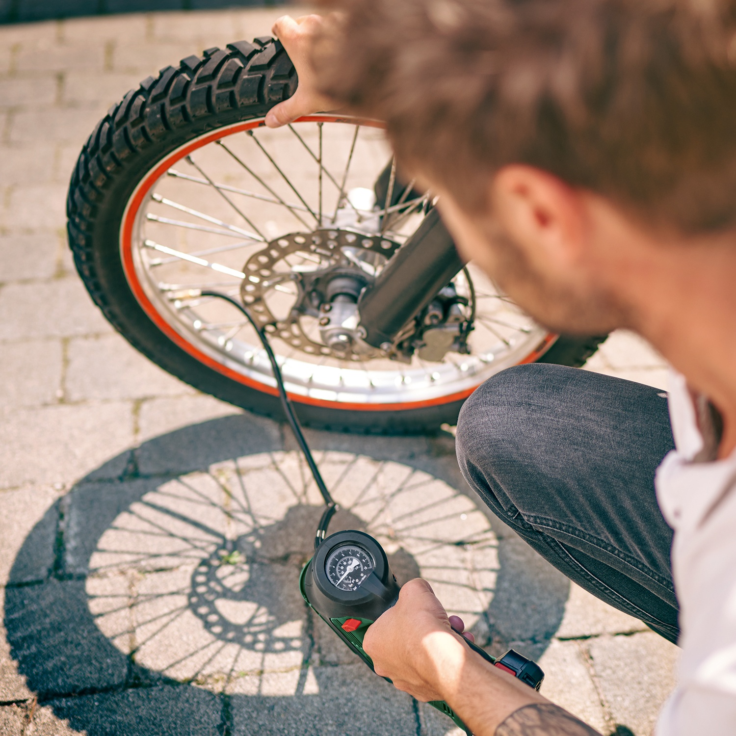 Una persona gonfia uno pneumatico di moto con un manometro. Lo pneumatico è nero con cerchio rosso. La persona indossa jeans grigi.