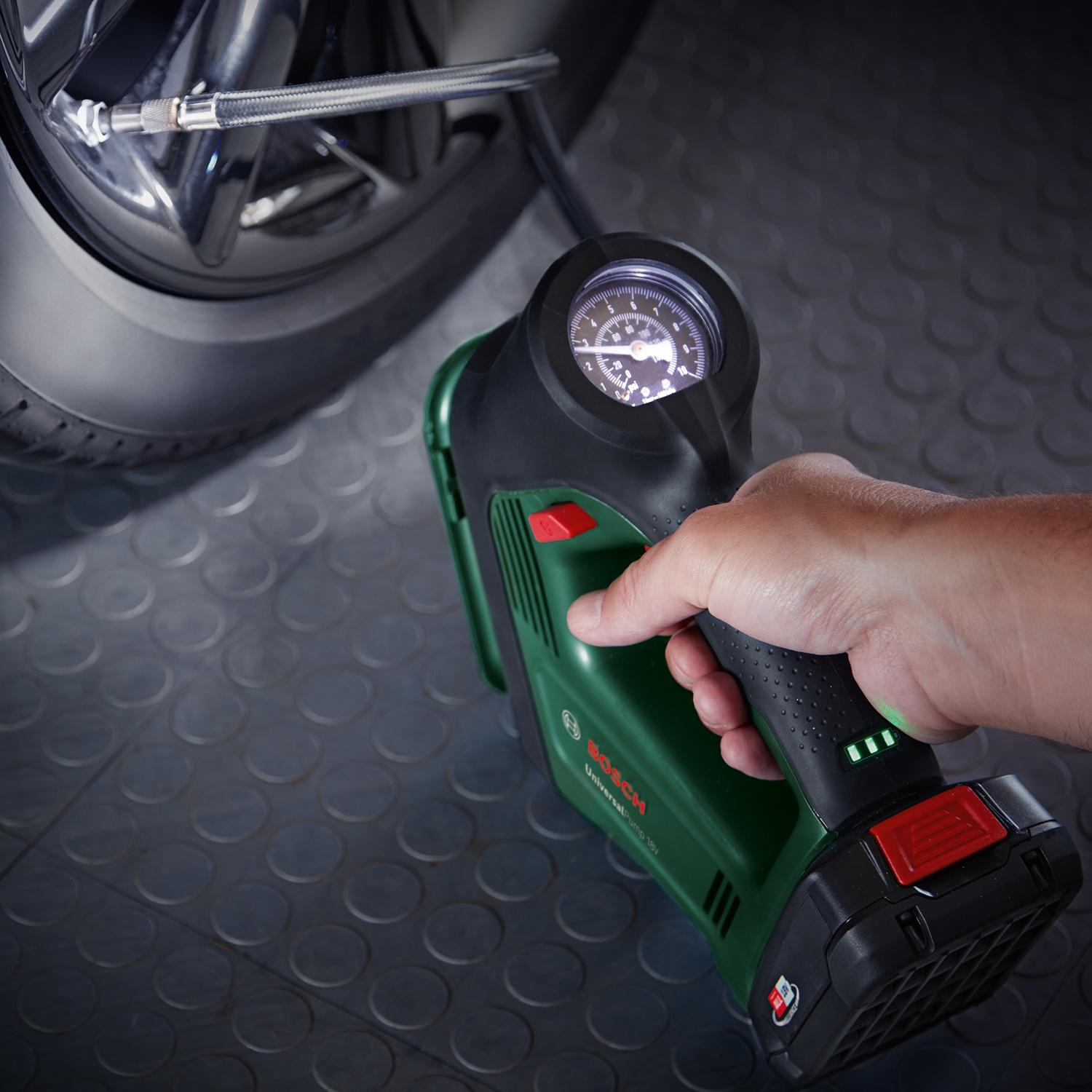 Una mano tiene un gonfiatore per pneumatici Bosch vicino a uno pneumatico per auto. Il manometro del gonfiatore è illuminato. Lo sfondo presenta un pavimento a motivi.