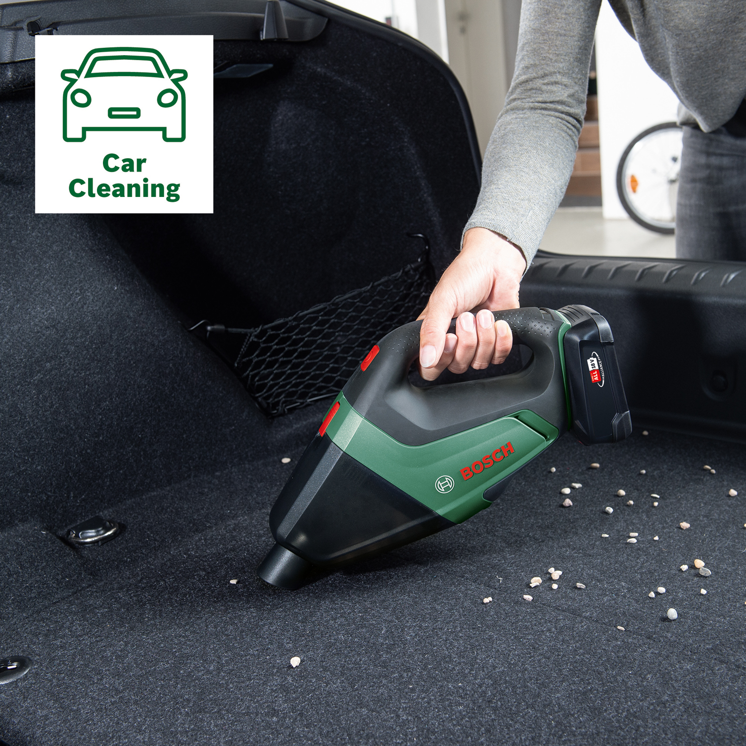 Una persona usa un aspirapolvere portatile Bosch nero e verde per pulire il bagagliaio di un'auto, con piccole pietre sul tappeto.