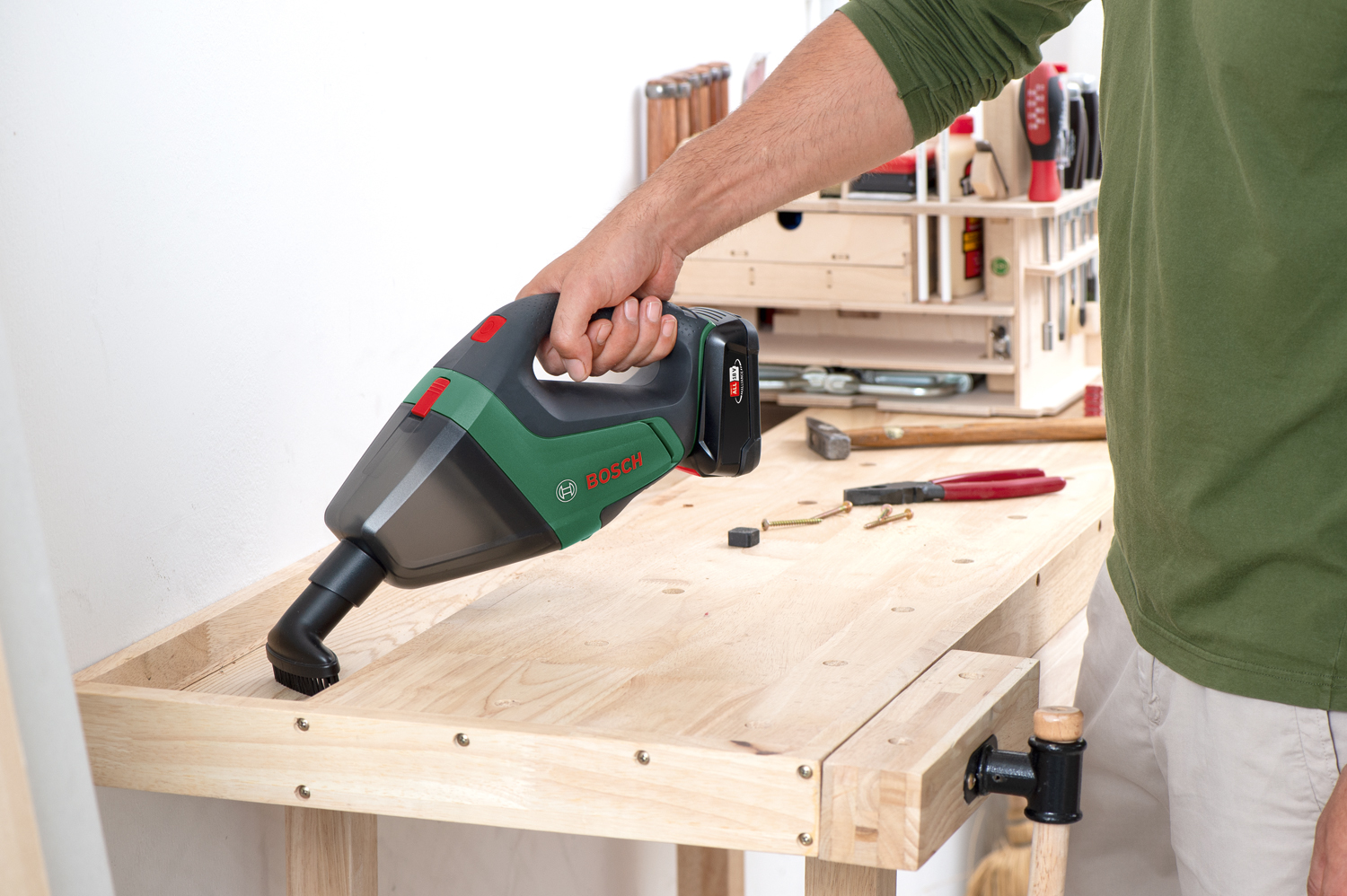 Una persona tiene in mano un aspirapolvere portatile Bosch sopra un banco da lavoro in legno. Utensili e forniture sono sul banco e sullo sfondo.