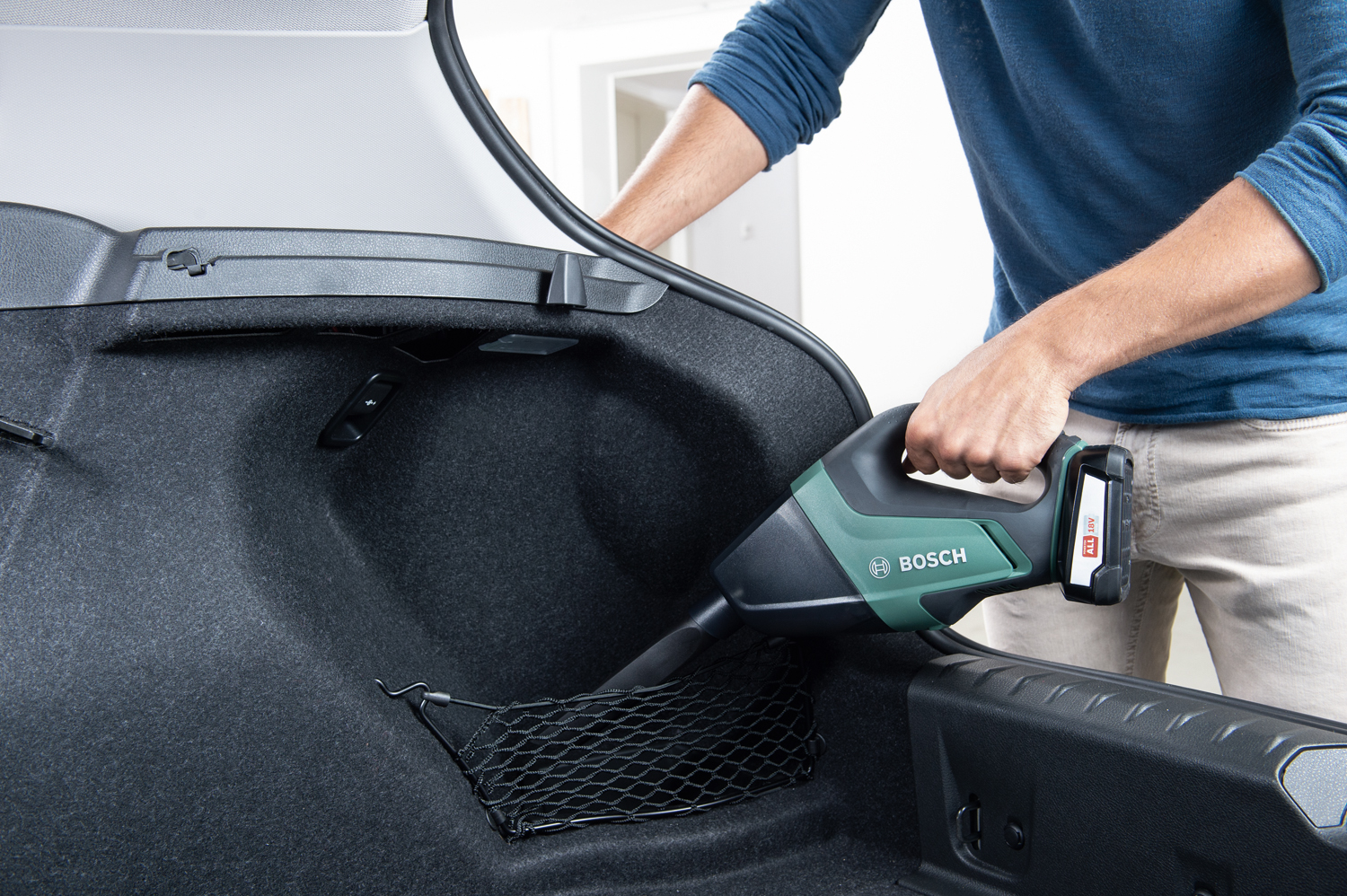 Una persona aspira il bagagliaio di un'auto con un aspirapolvere Bosch. Il bagagliaio è aperto e l'aspirapolvere è tenuto nella mano destra.