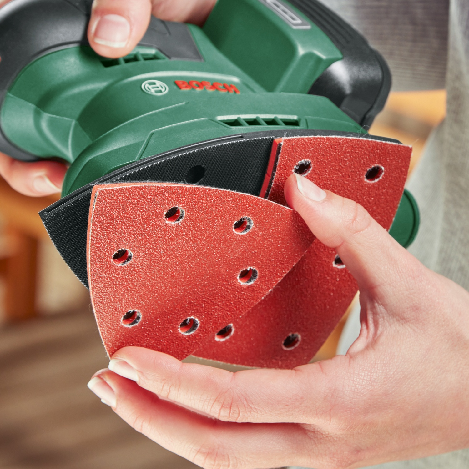 Mani che tengono una levigatrice Bosch verde con tamponi rossi. Focus sui fori e dettagli. Sfondo sfocato.