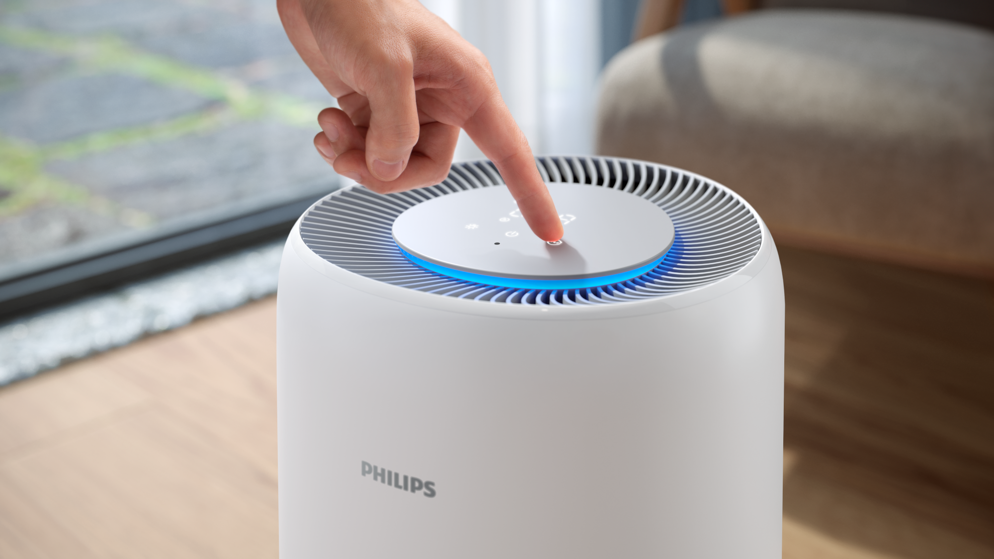 Egy kéz megnyomja a bekapcsológombot egy fehér Philips légtisztítón. Kék fénye van, és egy fa padlón van.