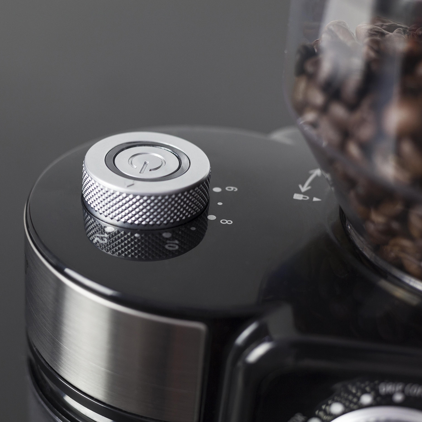 Zbliżenie na młynek do kawy. Srebrny przycisk zasilania na górze, czarny korpus i przezroczysty pojemnik z ziarnami kawy po prawej.