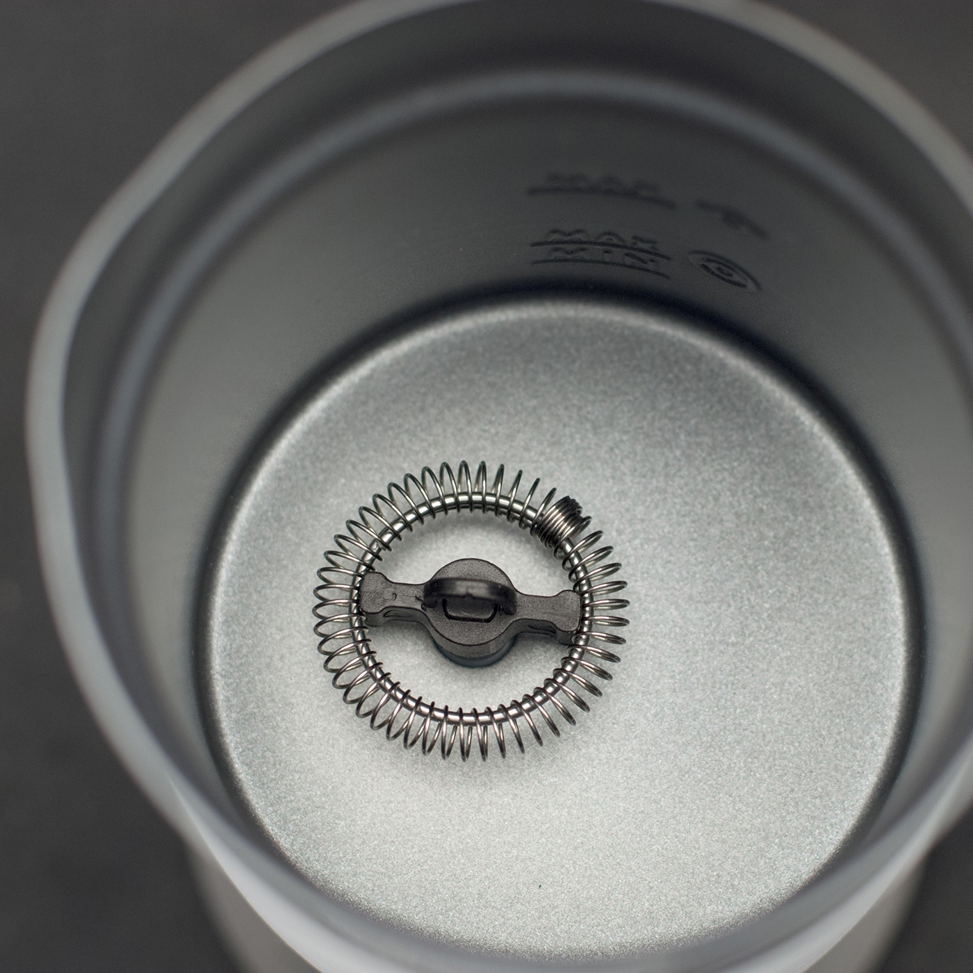 Zbliżenie spieniacza do mleka wewnątrz pojemnika. Spiralna metalowa trzepaczka jest wyśrodkowana. Pojemnik jest szary, z oznaczeniami MAX.