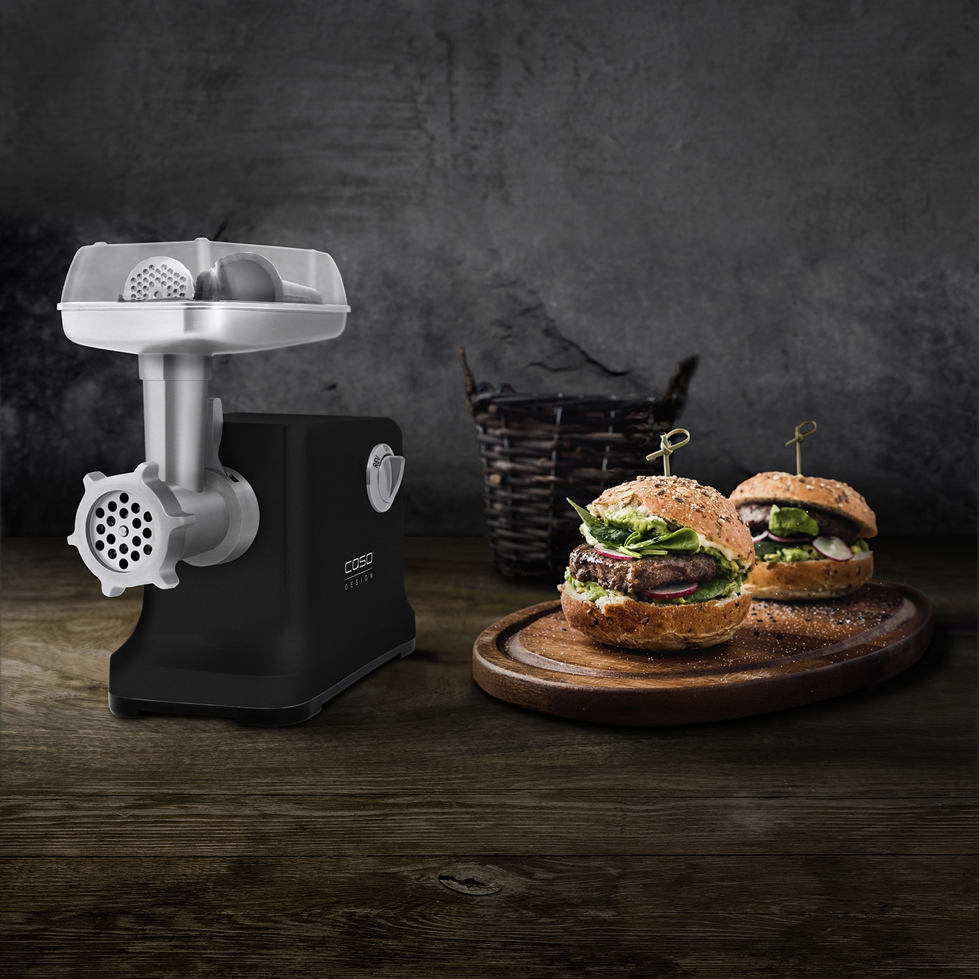 Czarna maszynka do mięsa stoi obok dwóch burgerów na drewnianej desce. Kosz w tle, na drewnianym stole.