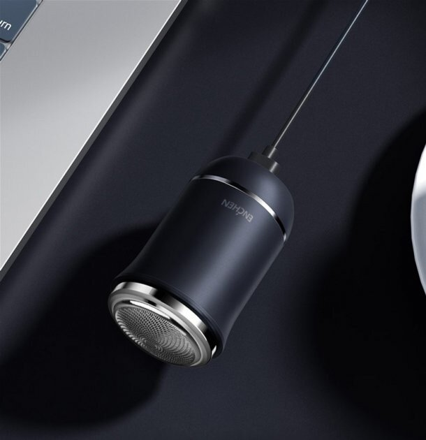 Czarna golarka elektryczna ze srebrną obwódką, połączona przewodem. Leży na ciemnej powierzchni w pobliżu laptopa i białego przedmiotu.