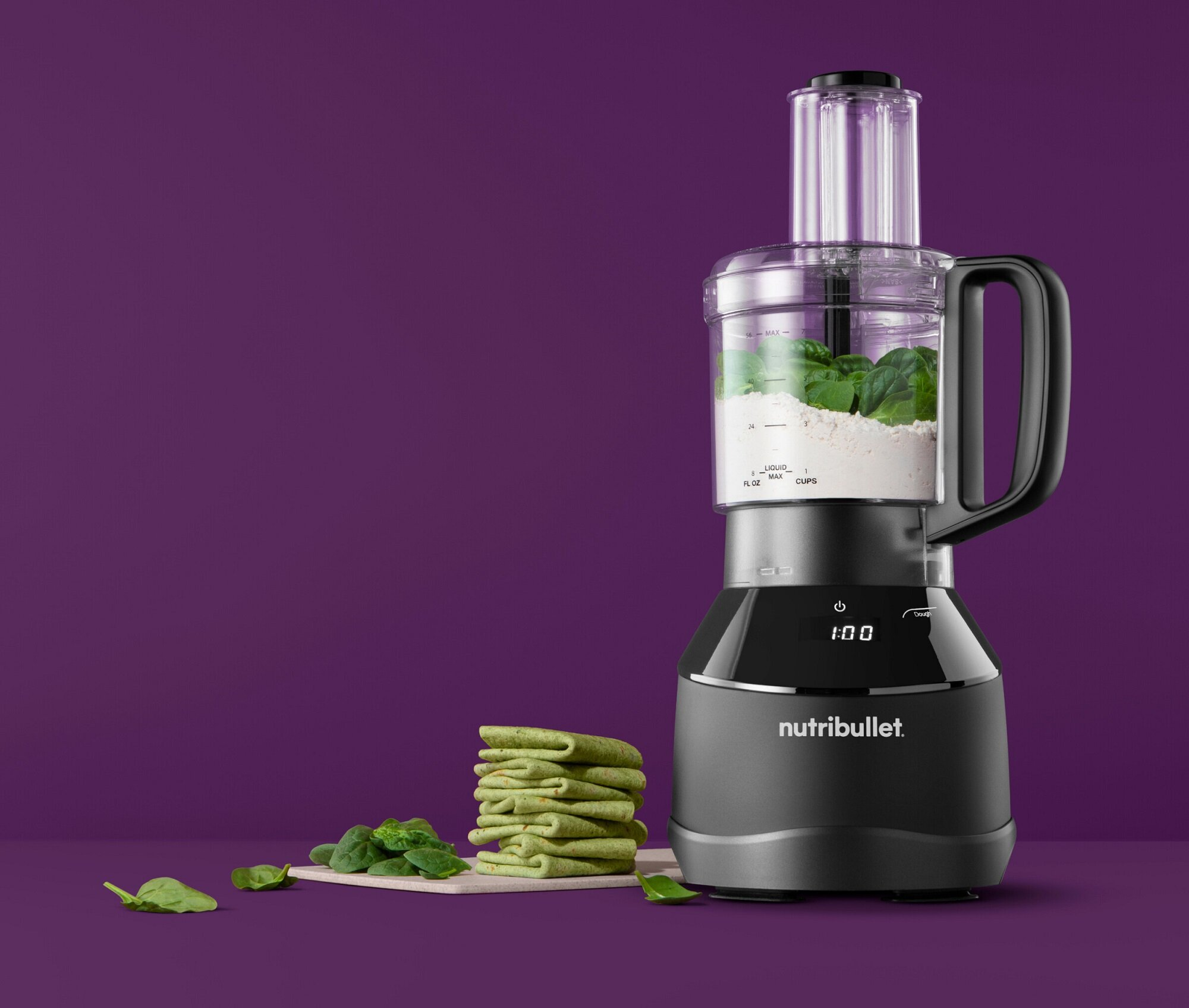 Czarny robot kuchenny Nutribullet na fioletowym tle, z tortillami, szpinakiem i składnikami w środku.