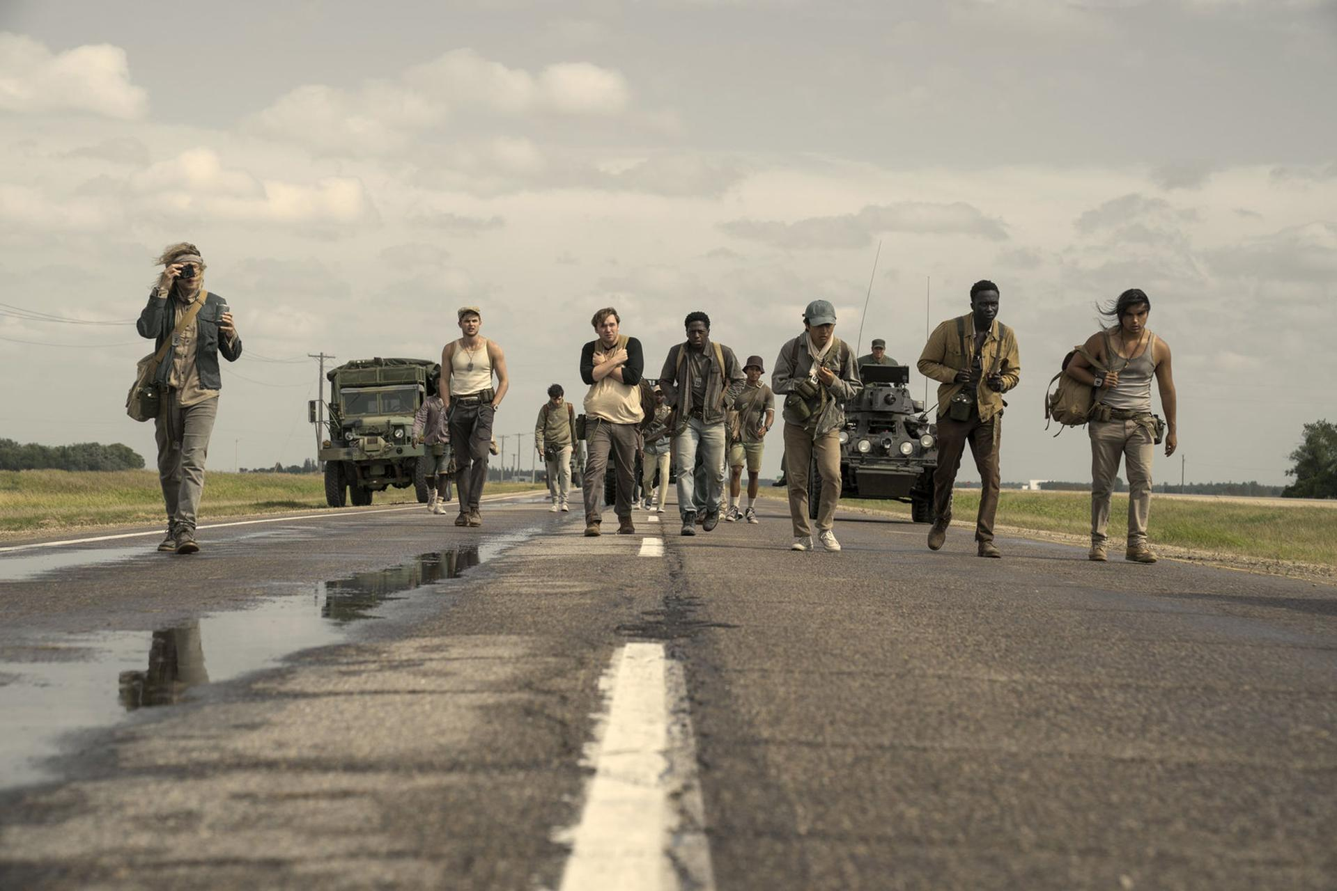 Gruppe von Personen, die auf einer Straße gehen, mit Militärfahrzeugen. Bewölkter Himmel und grüne Felder im Hintergrund.