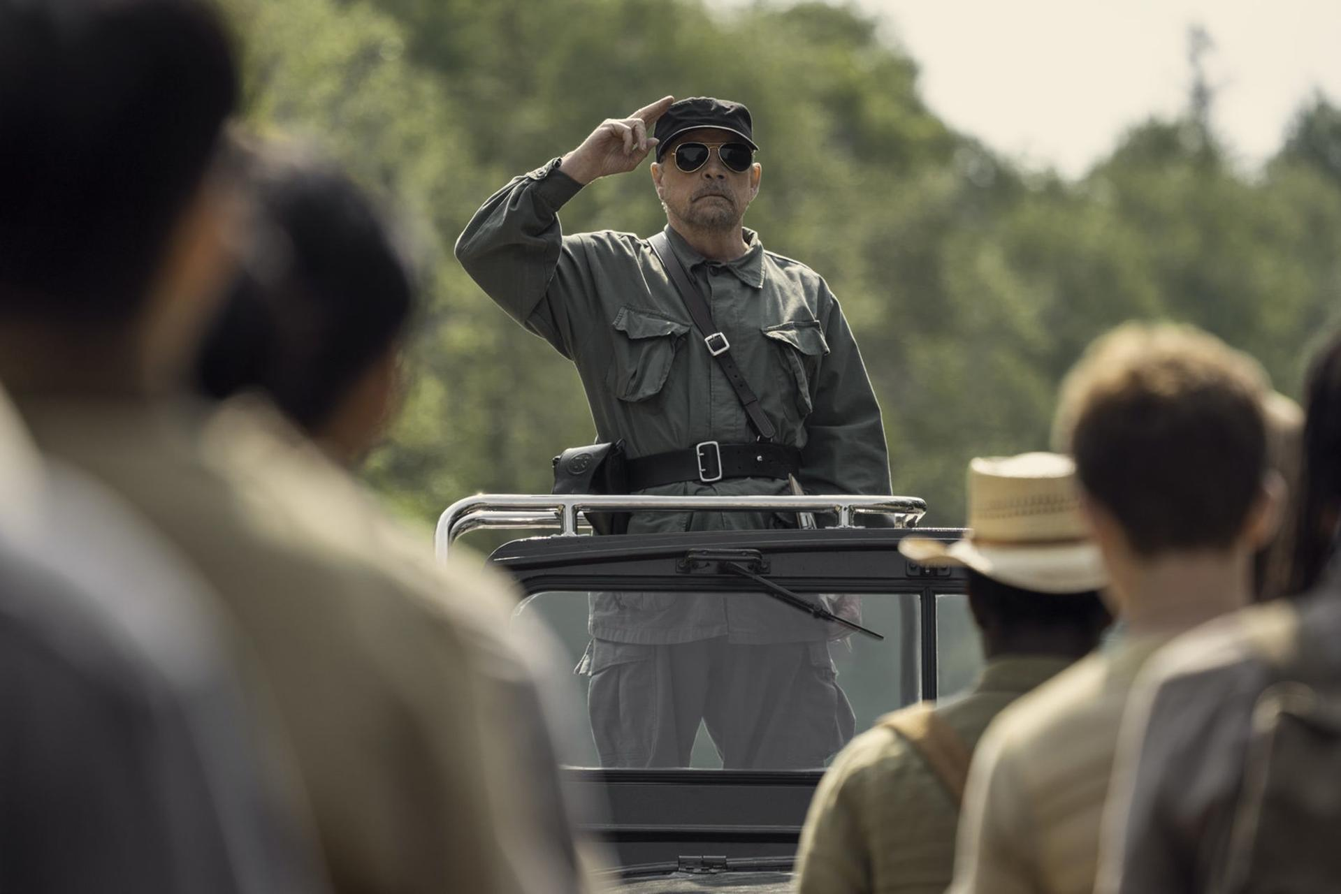 Ein Mann in Uniform salutiert von einem Fahrzeug aus. Er trägt eine Sonnenbrille und eine Mütze. Menschen sind im Vorder- und Hintergrund.