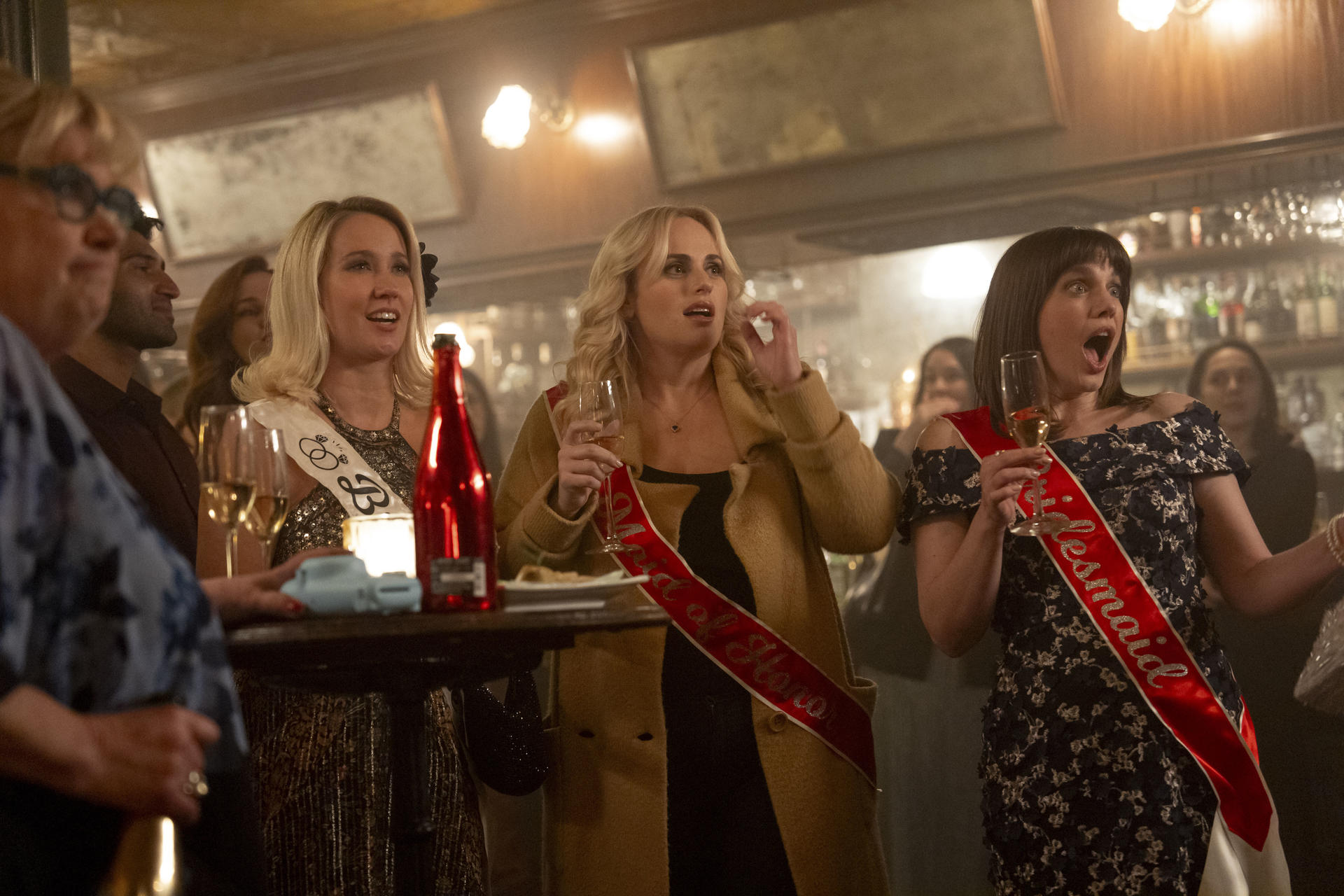 Drei Frauen in einer Bar, die Schärpen tragen. Eine sagt 'Braut', eine andere 'Trauzeugin' und die letzte 'Brautjungfer'. Sie halten Champagnergläser.
