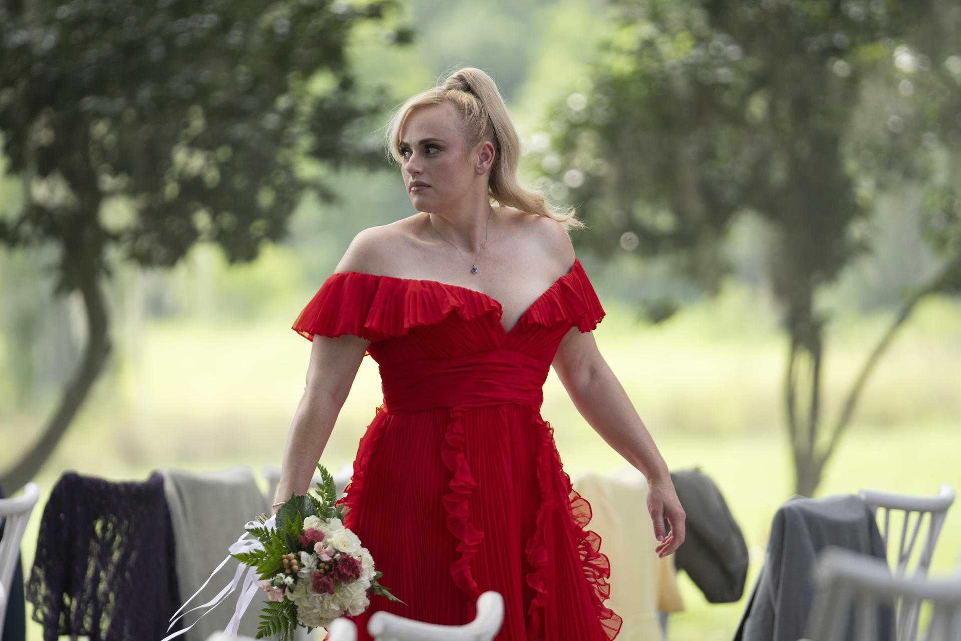 Frau in rotem Kleid mit Blumenstrauß. Sie ist im Freien mit Bäumen im Hintergrund. Tische und Stühle sind seitlich sichtbar.