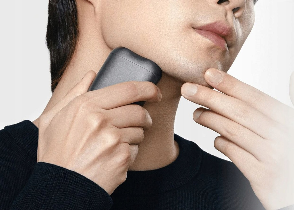 Mężczyzna goli policzek golarką elektryczną. Jego druga ręka prowadzi maszynkę. Ma na sobie czarny sweter.