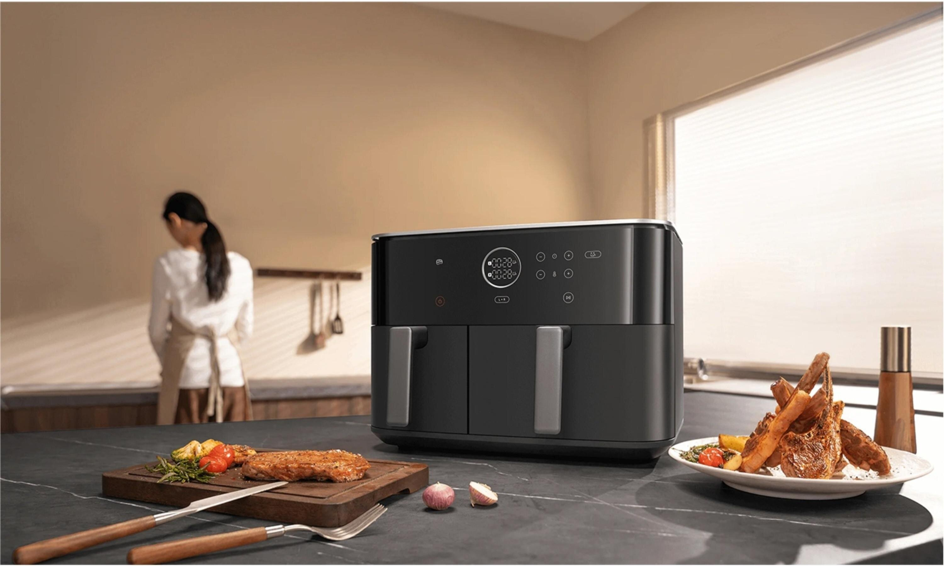 Frytkownica beztłuszczowa stoi na blacie z jedzeniem. Osoba w tle gotuje. Obecne są kotlety jagnięce i stek.