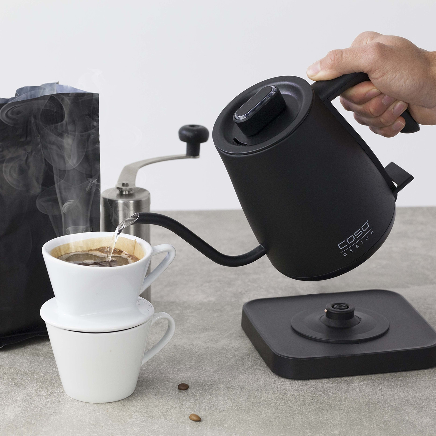 Osoba wlewa gorącą wodę z czarnego czajnika do filtra do kawy. W tle znajduje się młynek do kawy i worek z ziarnami kawy.