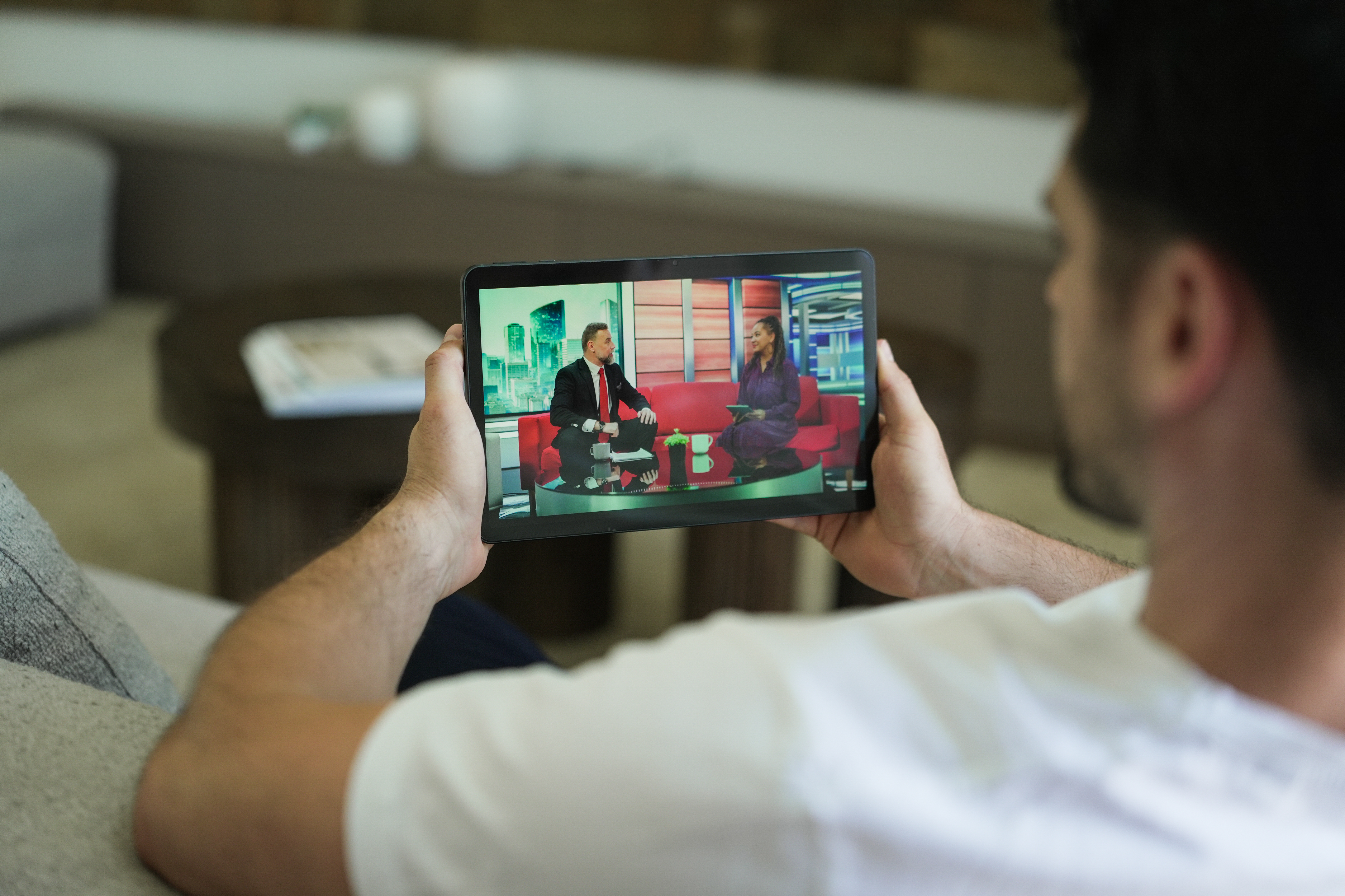 Ein Mann hält ein Tablet mit einem TV-Interview. Er sitzt auf einem Sofa, Hintergrund unscharf.