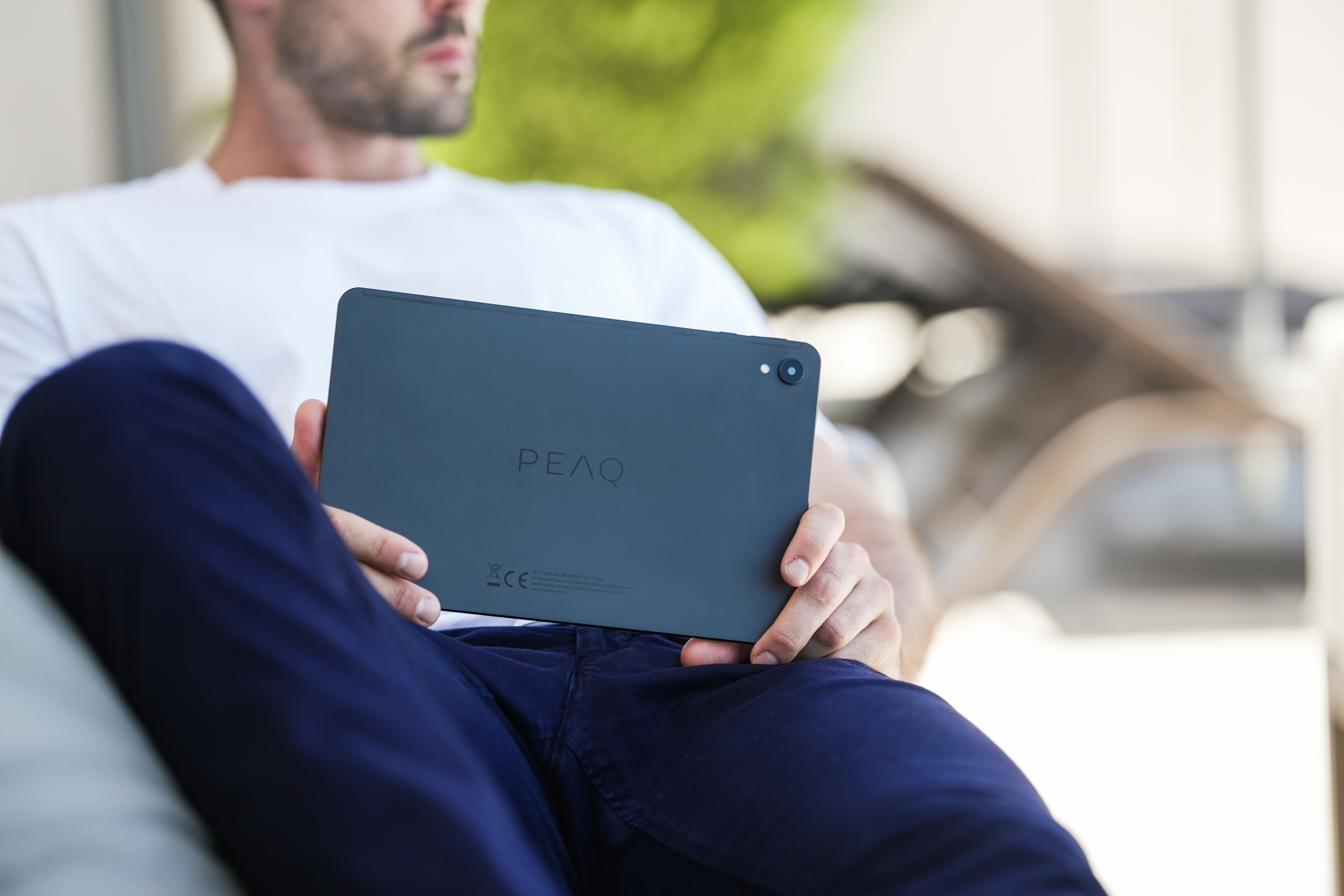 Ein Mann hält ein Tablet, die Rückseite ist sichtbar, mit der Marke 'PEAQ'. Er trägt ein weißes Hemd und blaue Hose.