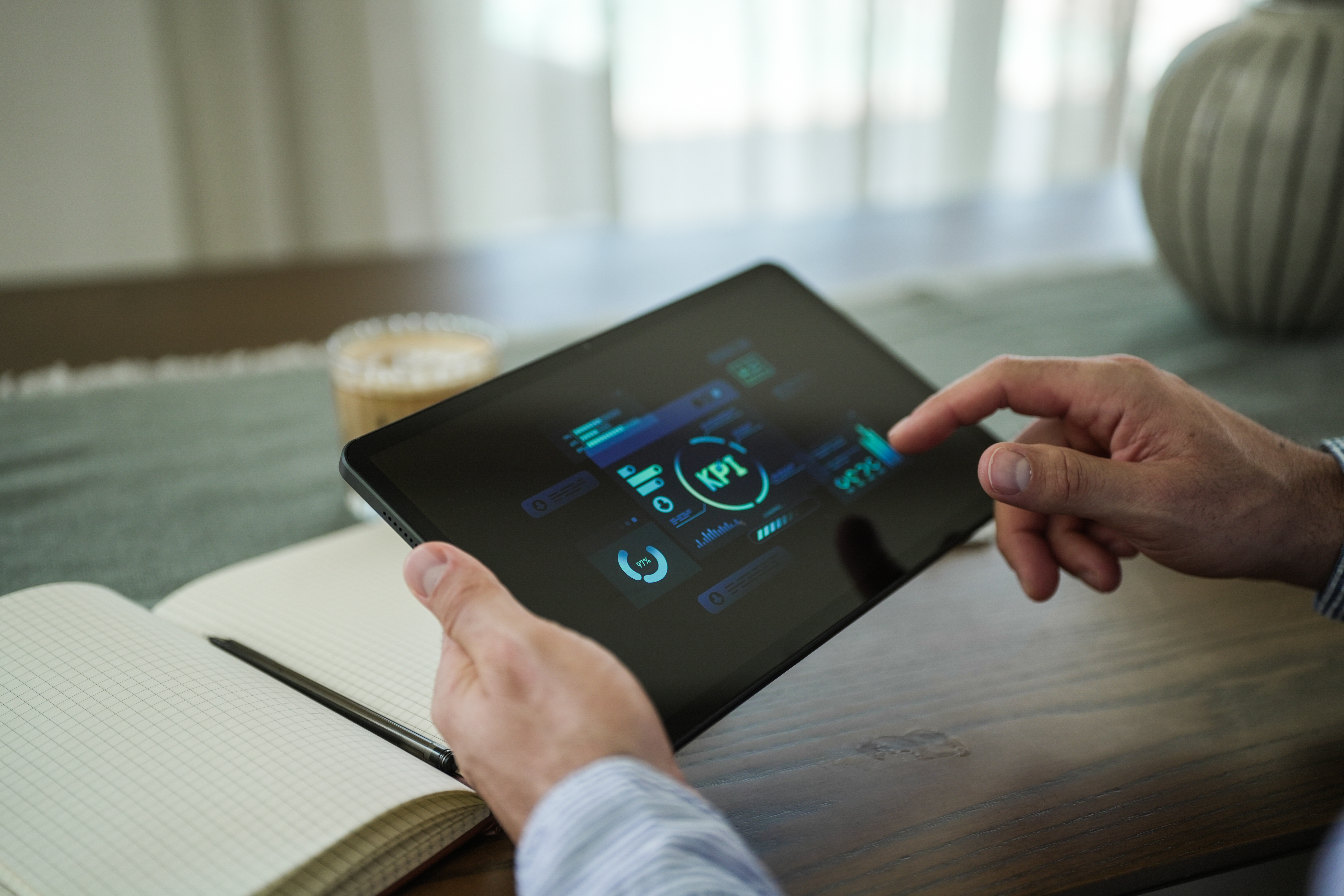 Eine Person benutzt ein Tablet, das ein KPI-Dashboard anzeigt. Ein Notizbuch und eine Kaffeetasse liegen auf dem Tisch. Die Person berührt den Bildschirm.
