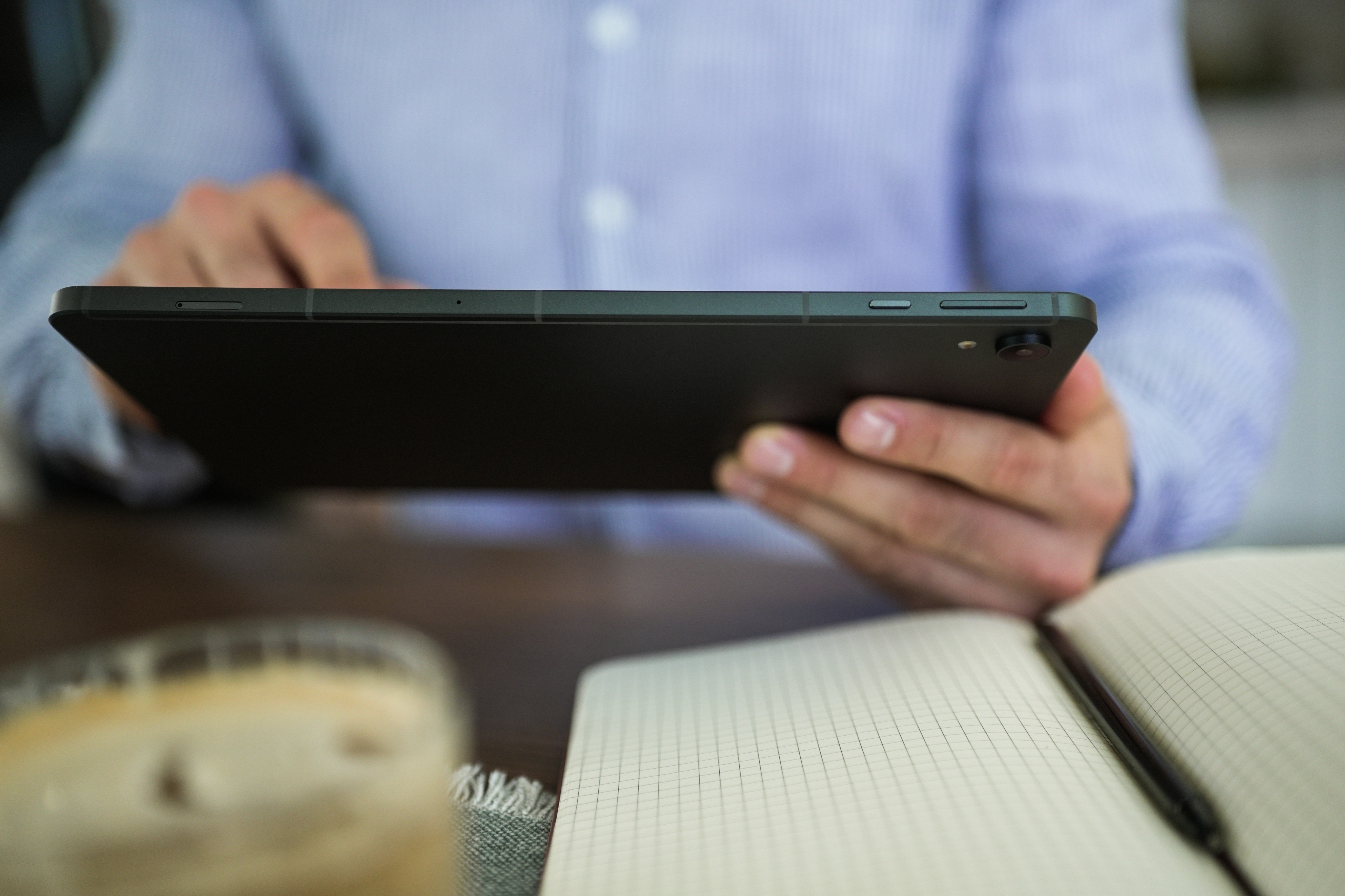 Eine Person hält ein schwarzes Tablet über einem Notizbuch mit kariertem Papier. Im Vordergrund ist Kaffee.