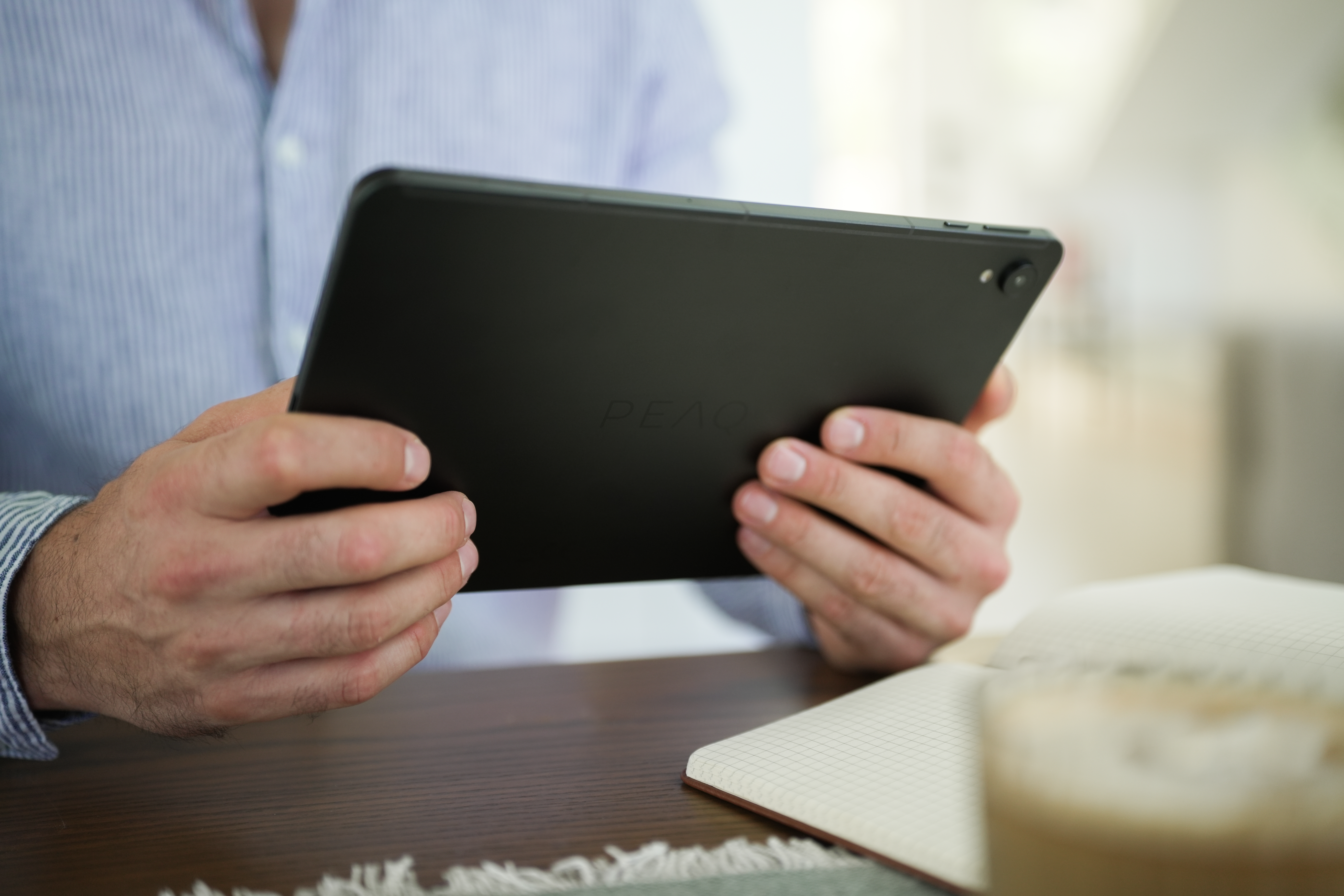 Mann hält ein schwarzes Tablet, im Hintergrund ein Notizblock und eine Kaffeetasse. Er trägt ein blaues Hemd.