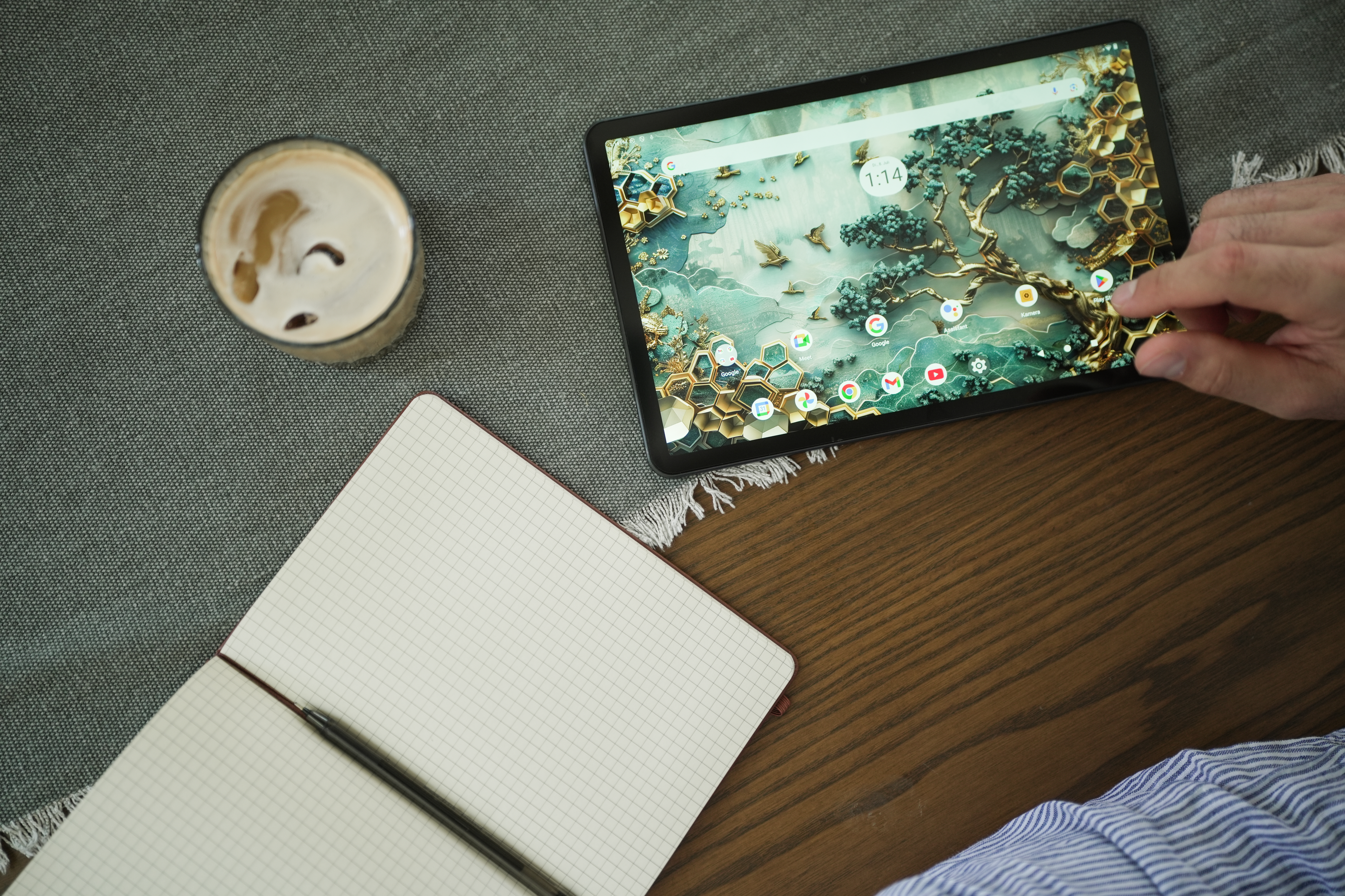 Ein Tablet, Kaffee und Notizbuch auf einem Holztisch. Eine Hand berührt den Tablet-Bildschirm.