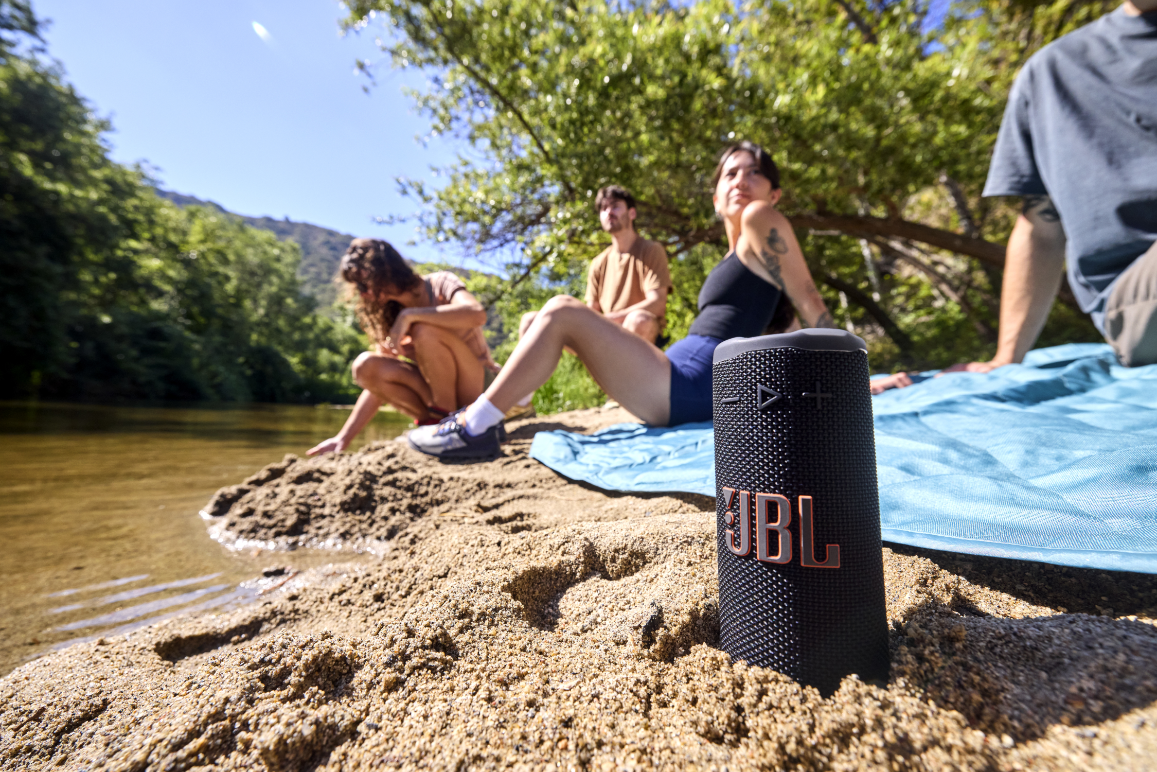 Un haut-parleur JBL noir sur du sable, près d'une rivière. Trois personnes sont à l'arrière-plan, assises. Des arbres et un ciel bleu sont visibles.
