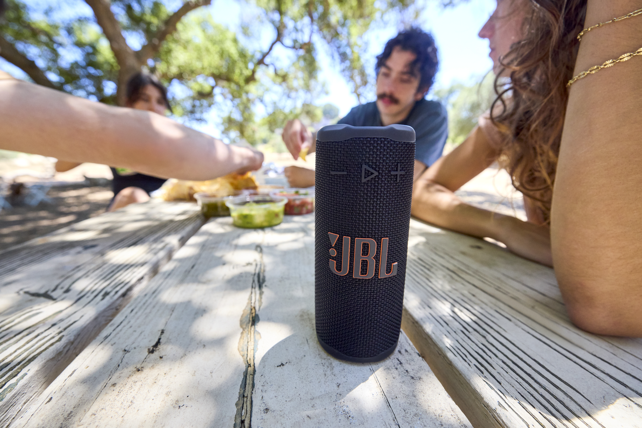 Une scène de pique-nique avec un haut-parleur JBL, des gens qui mangent et de la nourriture sur une table en bois. Il y a des arbres en arrière-plan.