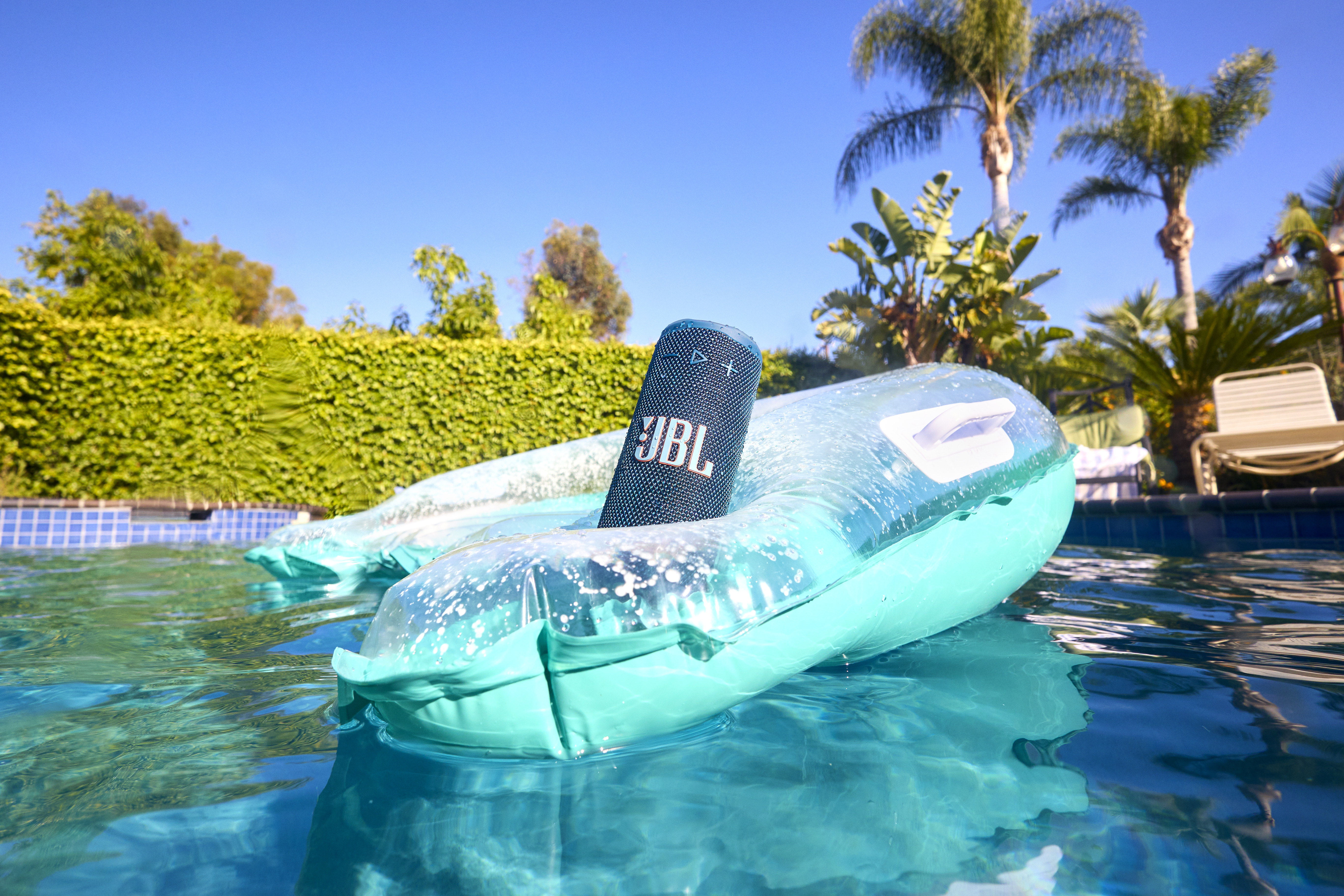 Un haut-parleur JBL flotte dans une piscine sur un gonflable. Des palmiers et de la verdure sont en arrière-plan sous un ciel bleu clair.