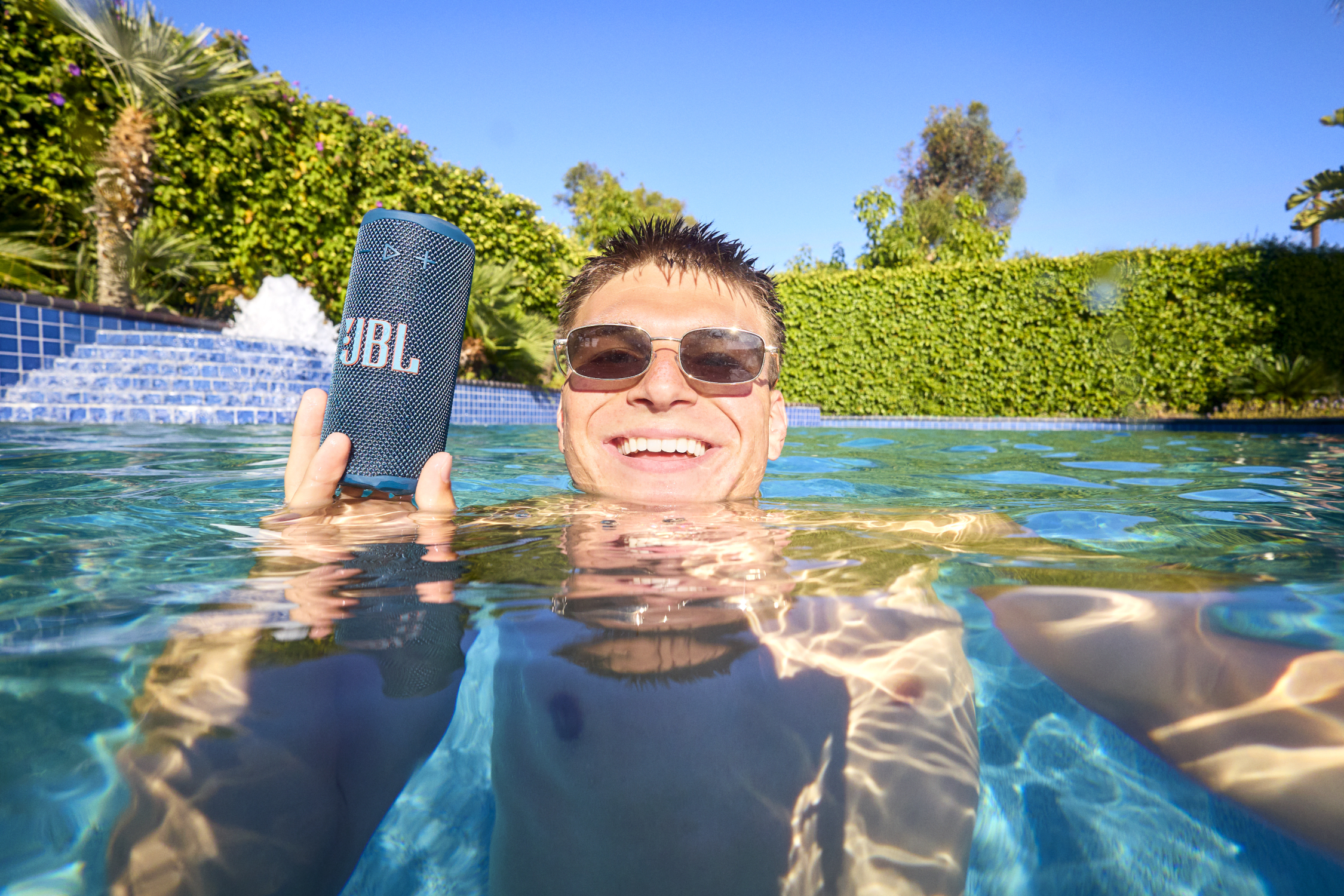 Un homme dans la piscine tient un haut-parleur JBL, souriant à la caméra. Le fond présente une fontaine, de la verdure et un ciel bleu.