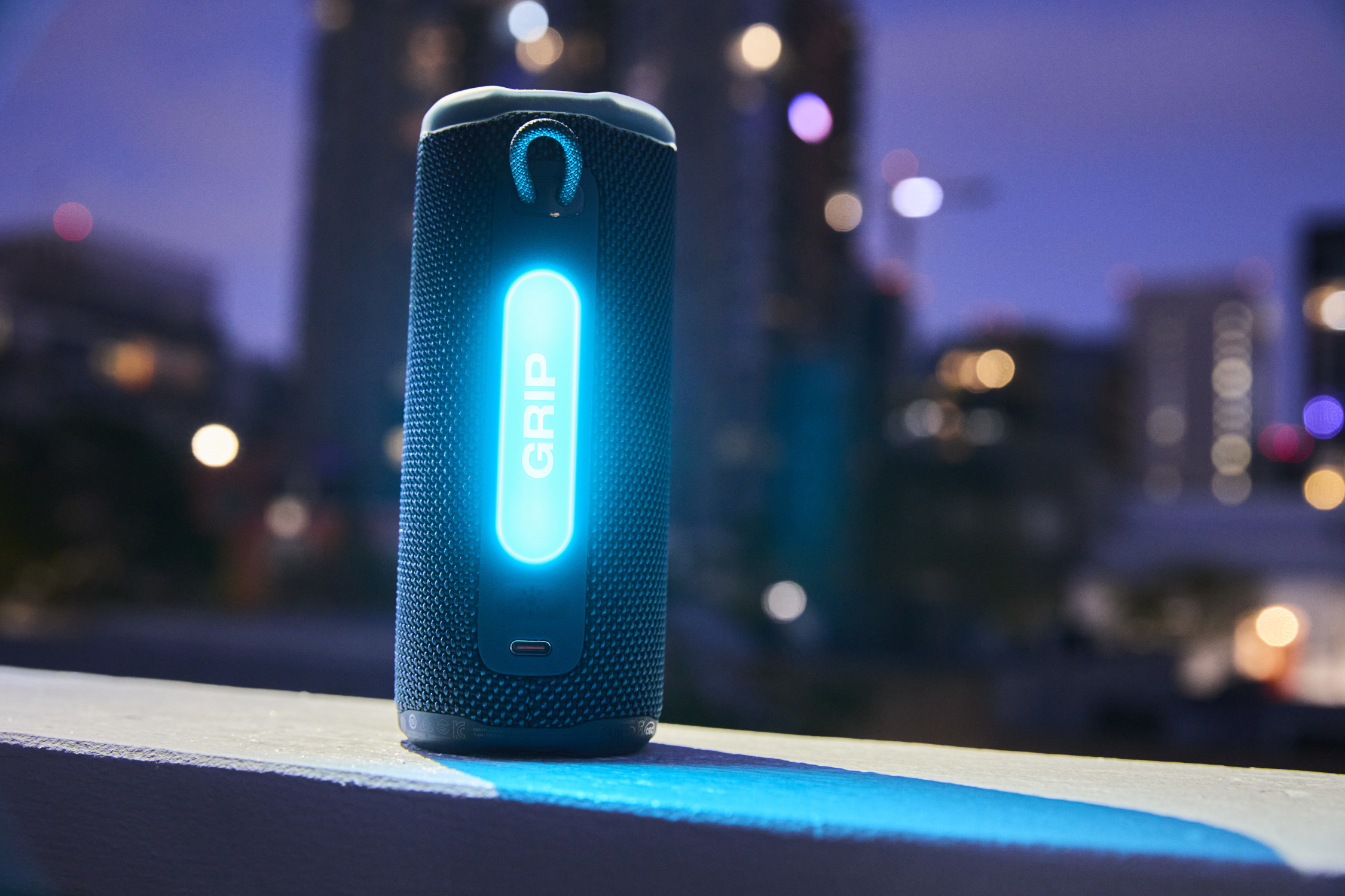 Un haut-parleur bleu avec le texte 'GRIP' lumineux sur un balcon. Lumières de la ville en arrière-plan flou au crépuscule.