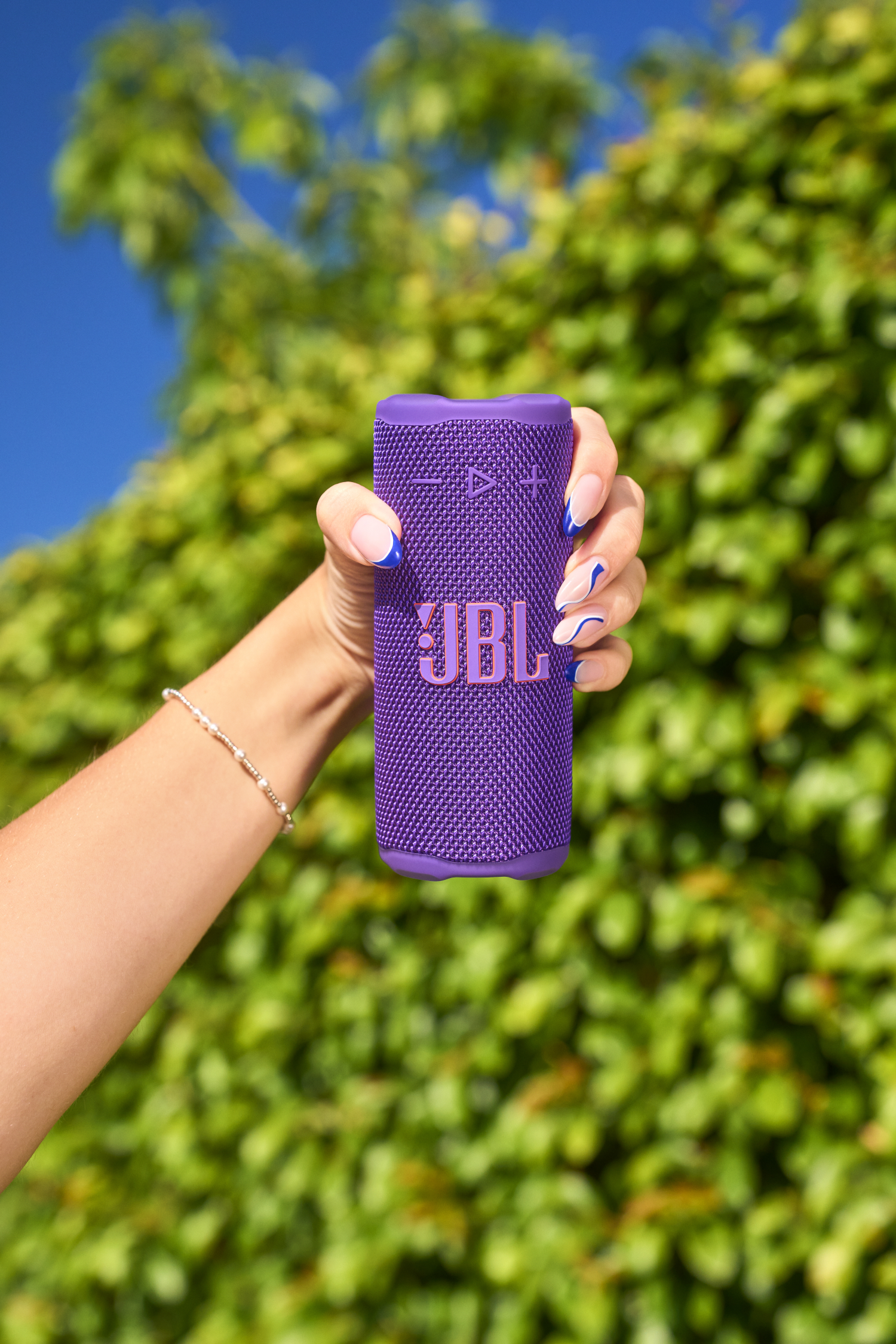Une main tient un haut-parleur JBL violet. Le fond montre des feuilles vertes et un ciel bleu.