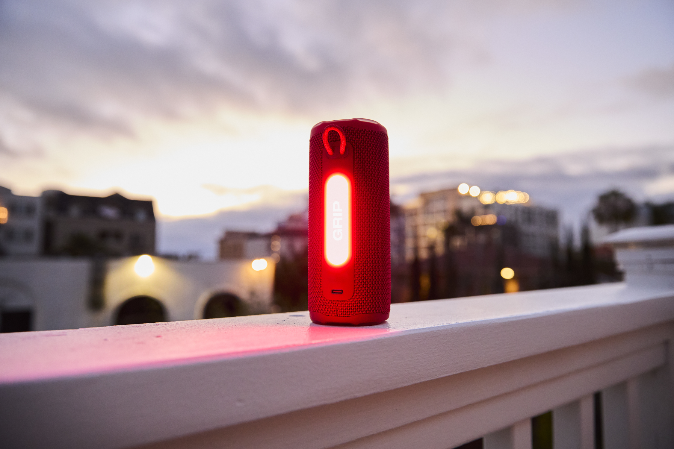 Un haut-parleur rouge avec une bande blanche brillante est posé sur une balustrade blanche au crépuscule. Bâtiments et ciel nuageux en arrière-plan.