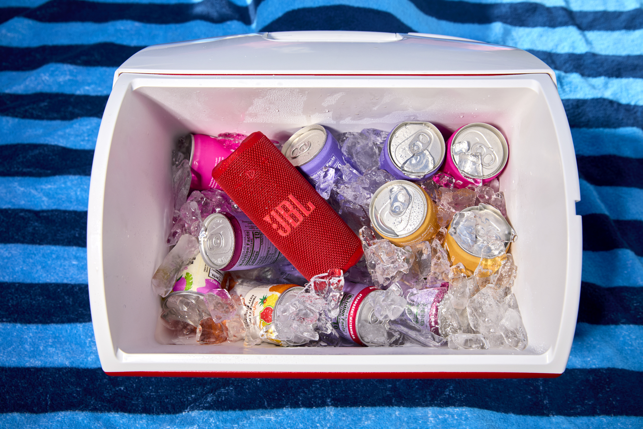Une glacière sur une serviette bleue à rayures contient de la glace, des canettes de jus et une enceinte JBL rouge.