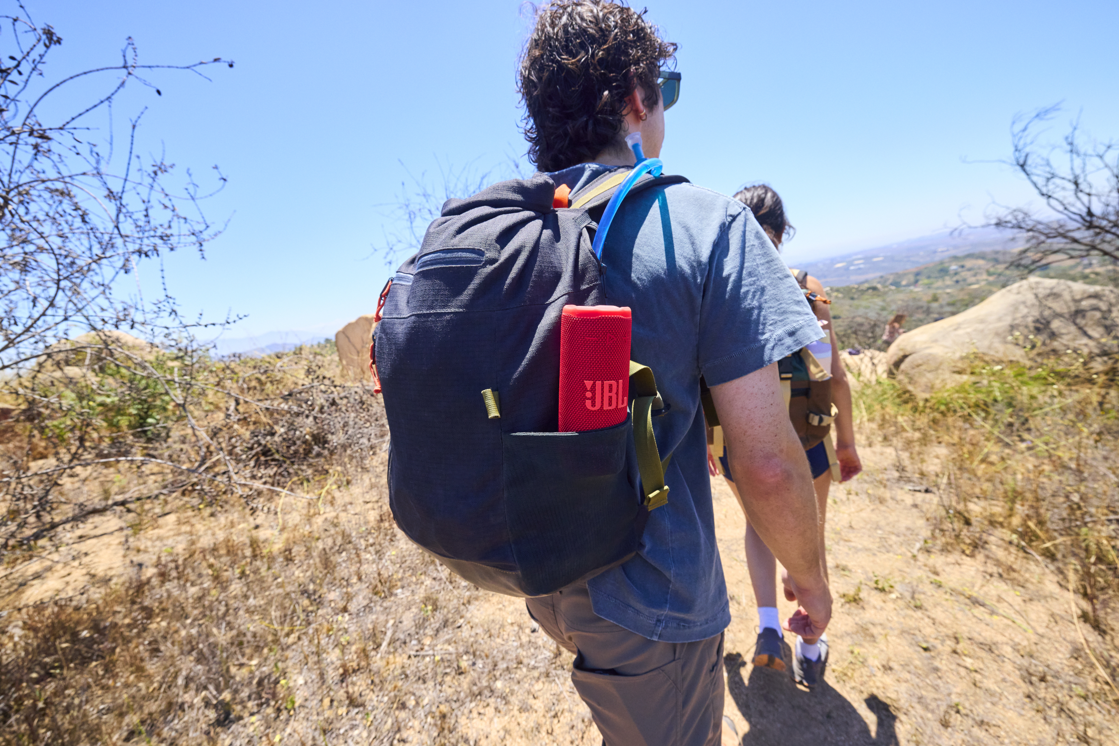 Personne faisant de la randonnée avec un sac à dos. Un haut-parleur JBL rouge est visible. Une autre personne marche devant. Ciel bleu clair et paysage sec en arrière-plan.