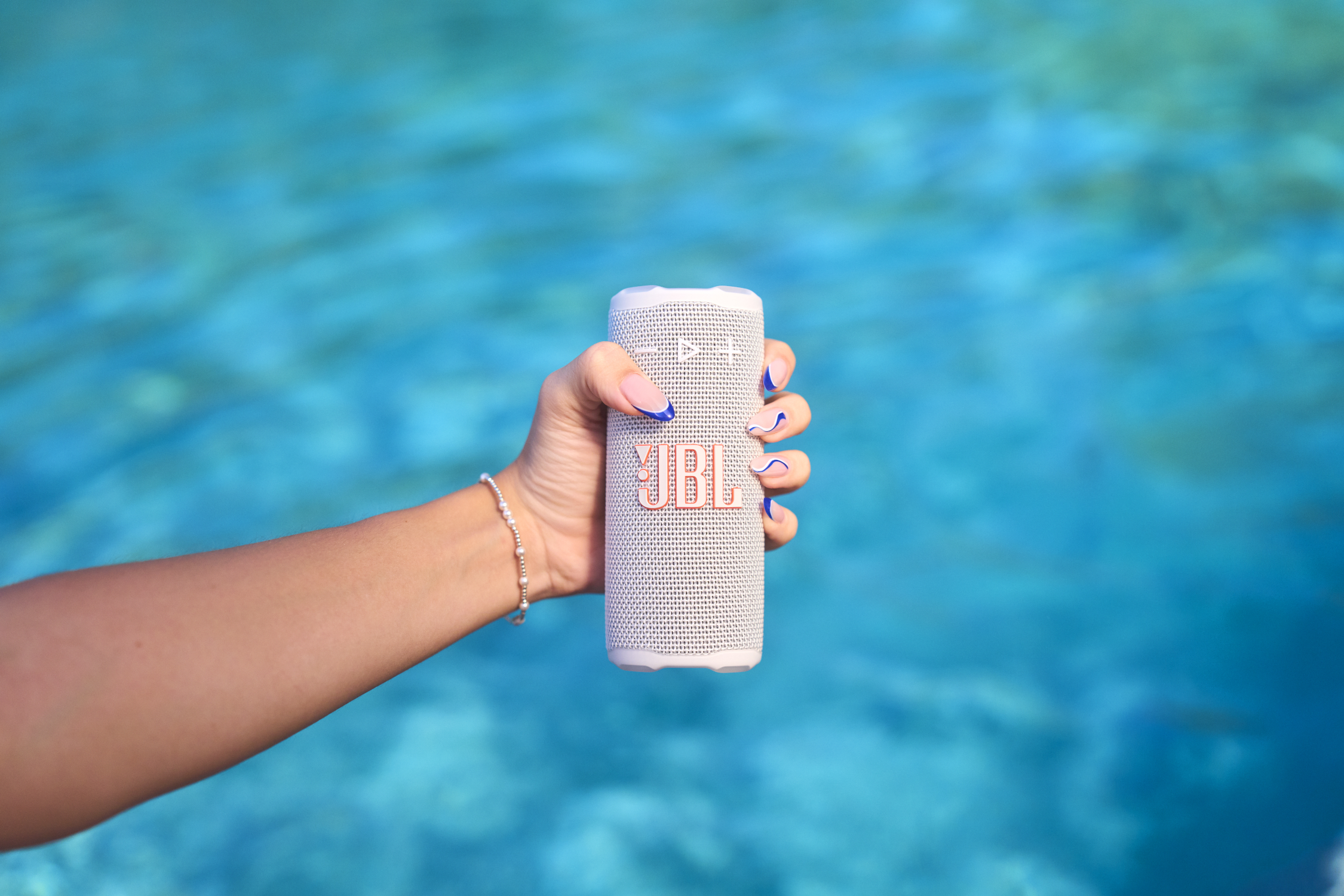 Main tenant un haut-parleur JBL blanc avec fond d'eau bleu. Ongles peints en bleu et blanc. Bracelet de perles au poignet.