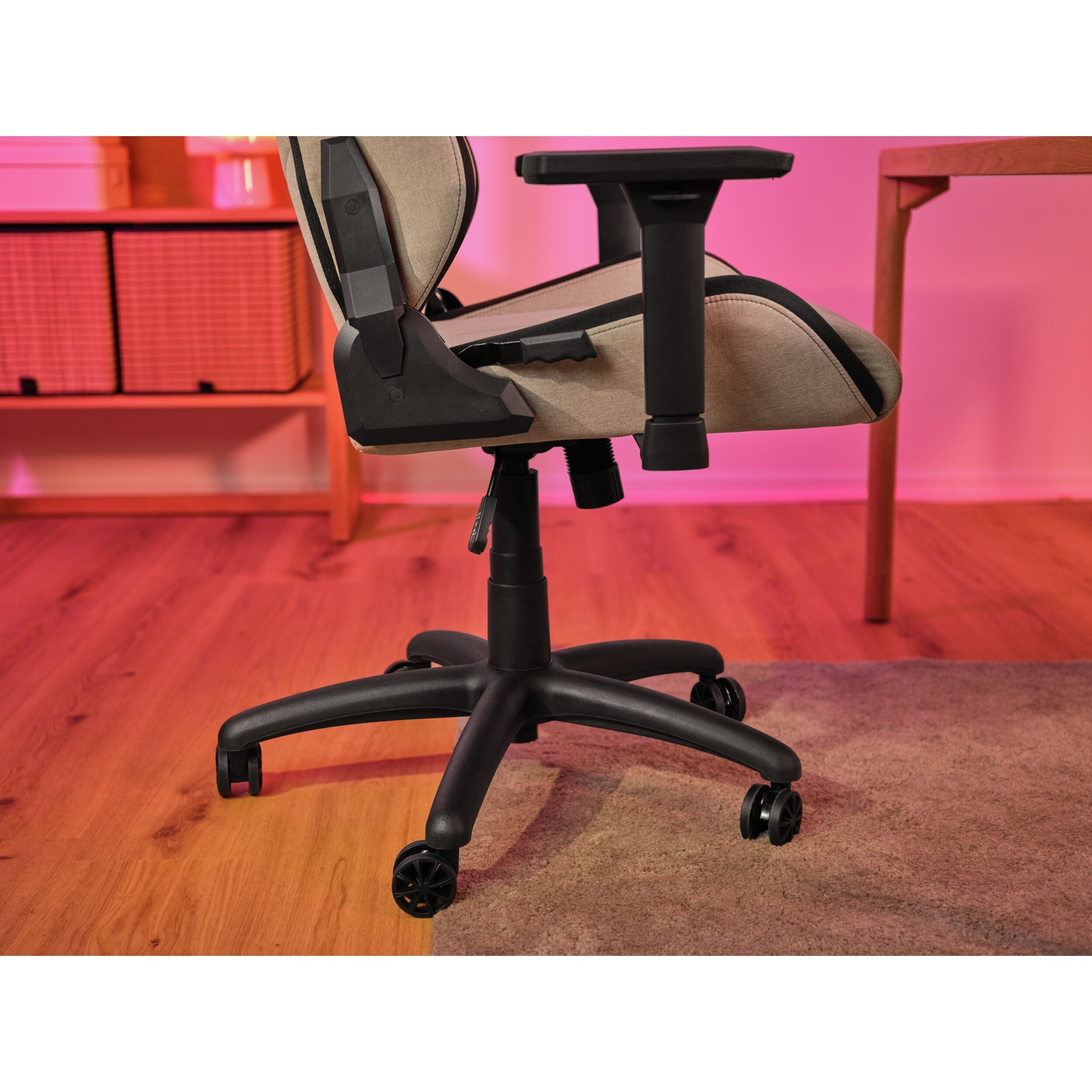 Una sedia da ufficio con seduta beige e dettagli neri. Ha base e ruote nere, su pavimento in legno con sfondo rosa.