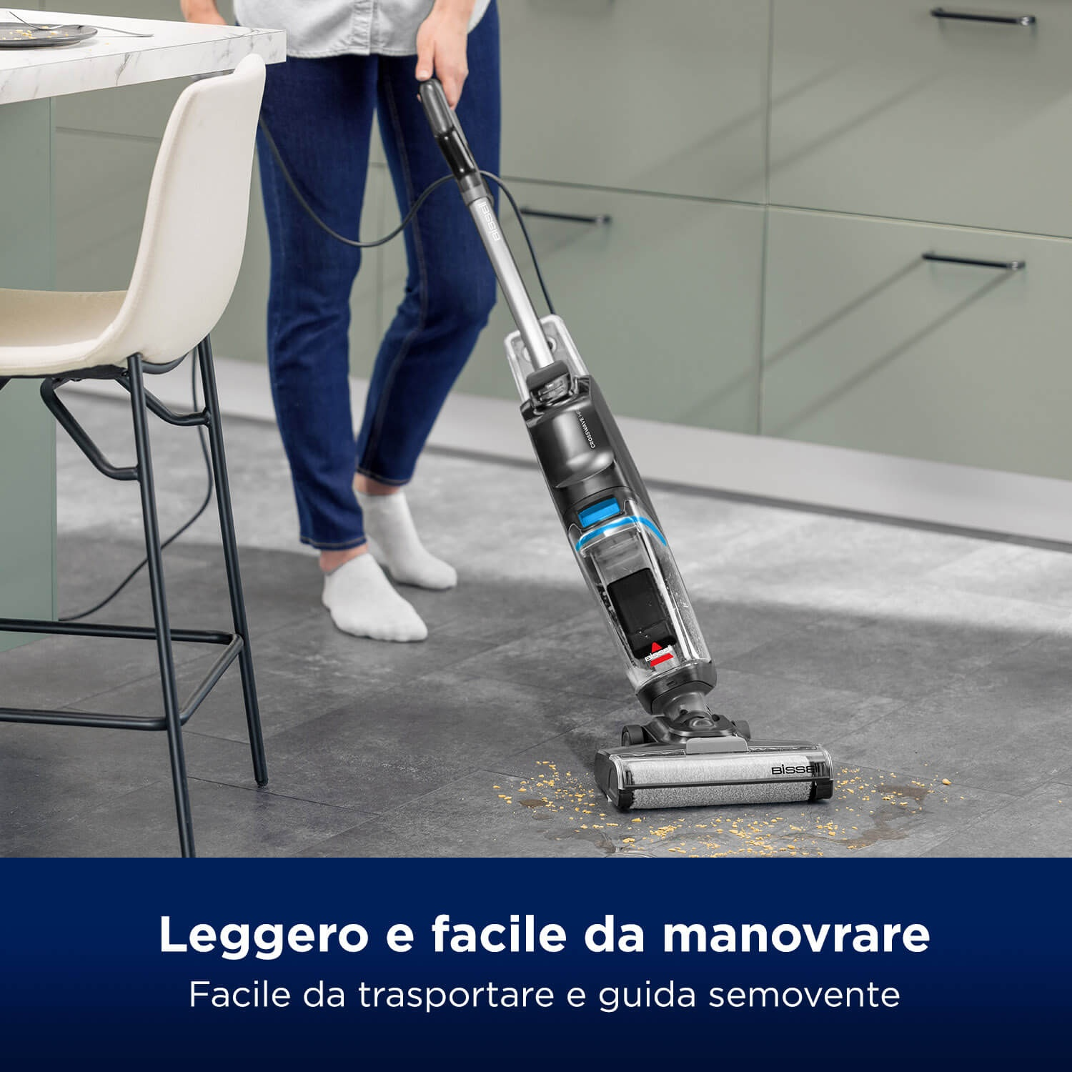 Persona aspira il pavimento della cucina. L'aspirapolvere è nero e argento. Il pavimento è grigio. La persona indossa jeans blu e calzini bianchi.