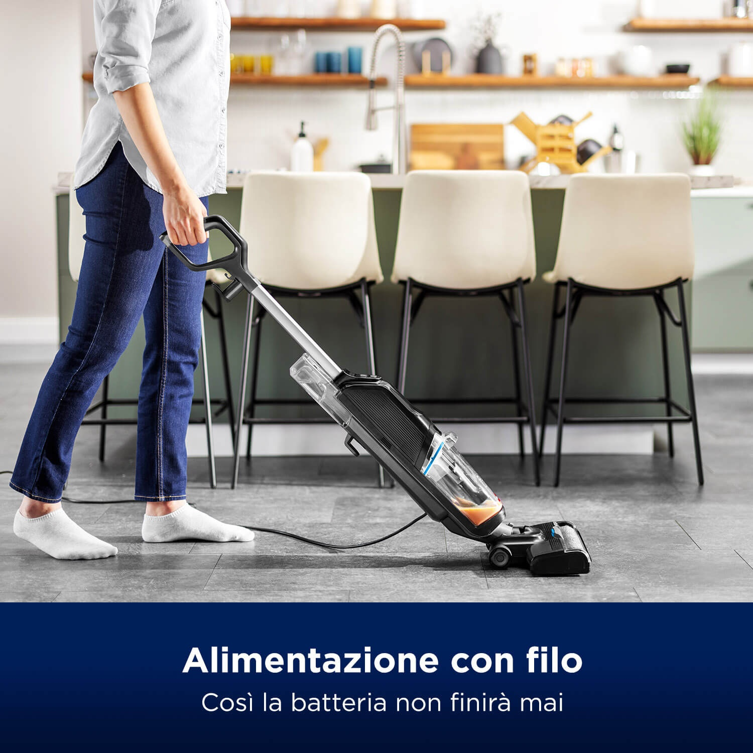 Una persona aspira un pavimento con un aspirapolvere nero. Lo sfondo include una cucina con sedie e un piano di lavoro.