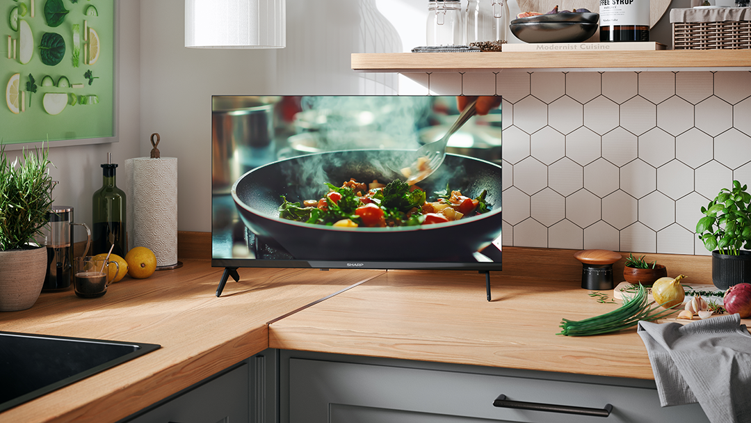 Egy konyhai jelenet, amelyen egy tévé látható, amin ételt főznek. Zöldségek és konyhai edények a pulton. Egy bekeretezett műalkotás lóg a falon.