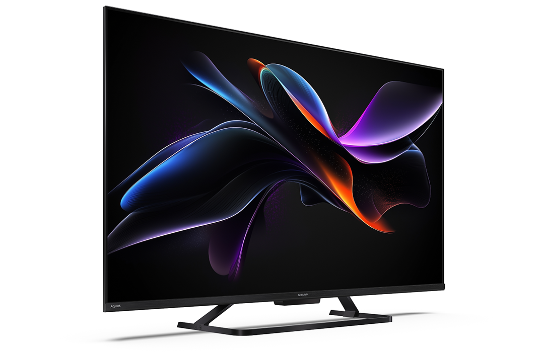 Egy nagy, modern Sharp Aquos TV. A képernyő színes, absztrakt formákat jelenít meg. Fekete állványon áll.