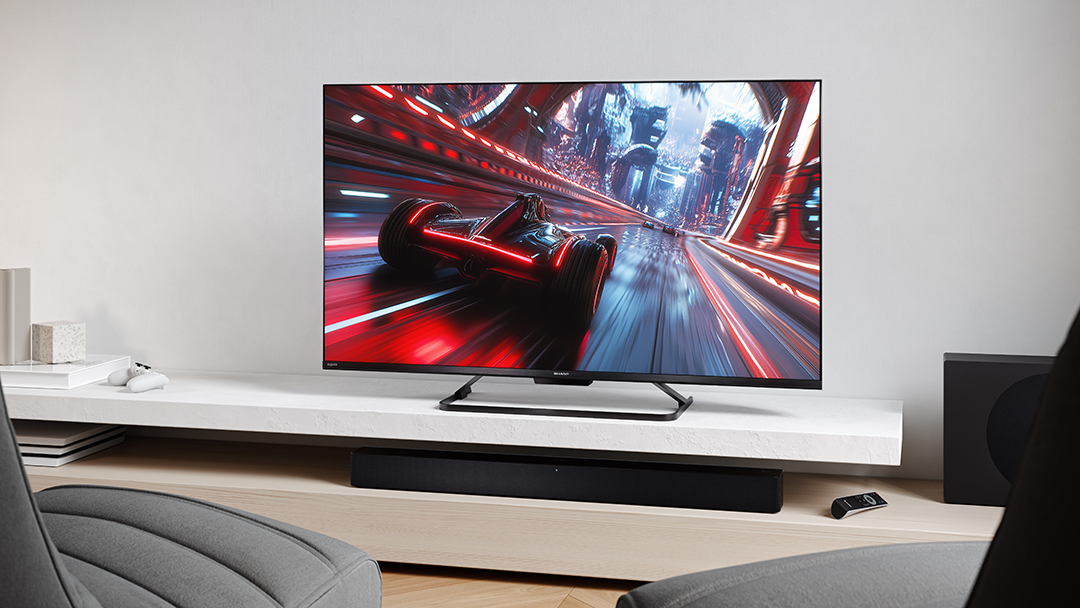 Egy TV-n egy versenyautó látható a pályán. Alatta egy soundbar fehér polcon, mellette egy távirányító.