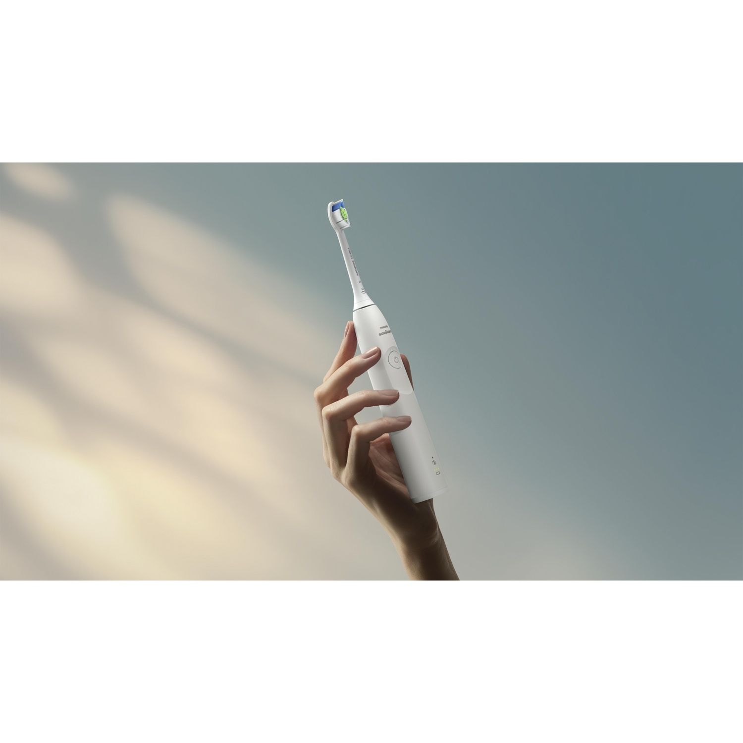 Una mano tiene uno spazzolino elettrico bianco su uno sfondo sfumato blu e giallo. La testina ha setole blu e verdi.